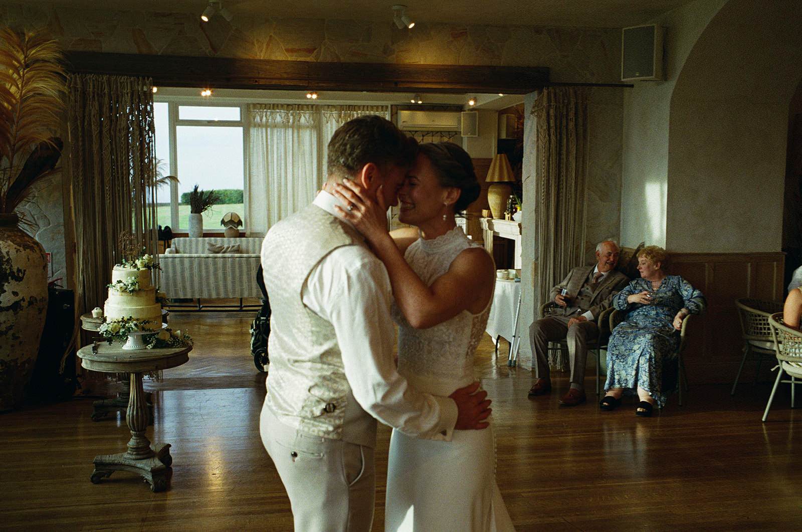 Bride and groom share a quiet first dance at their wedding reception as elderly guests watch from chairs.