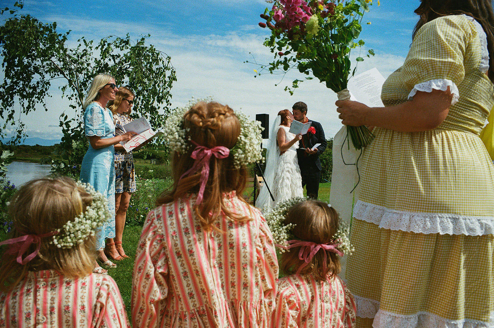 Outdoor wedding ceremony by river as couple read vows, children with flowers watching.