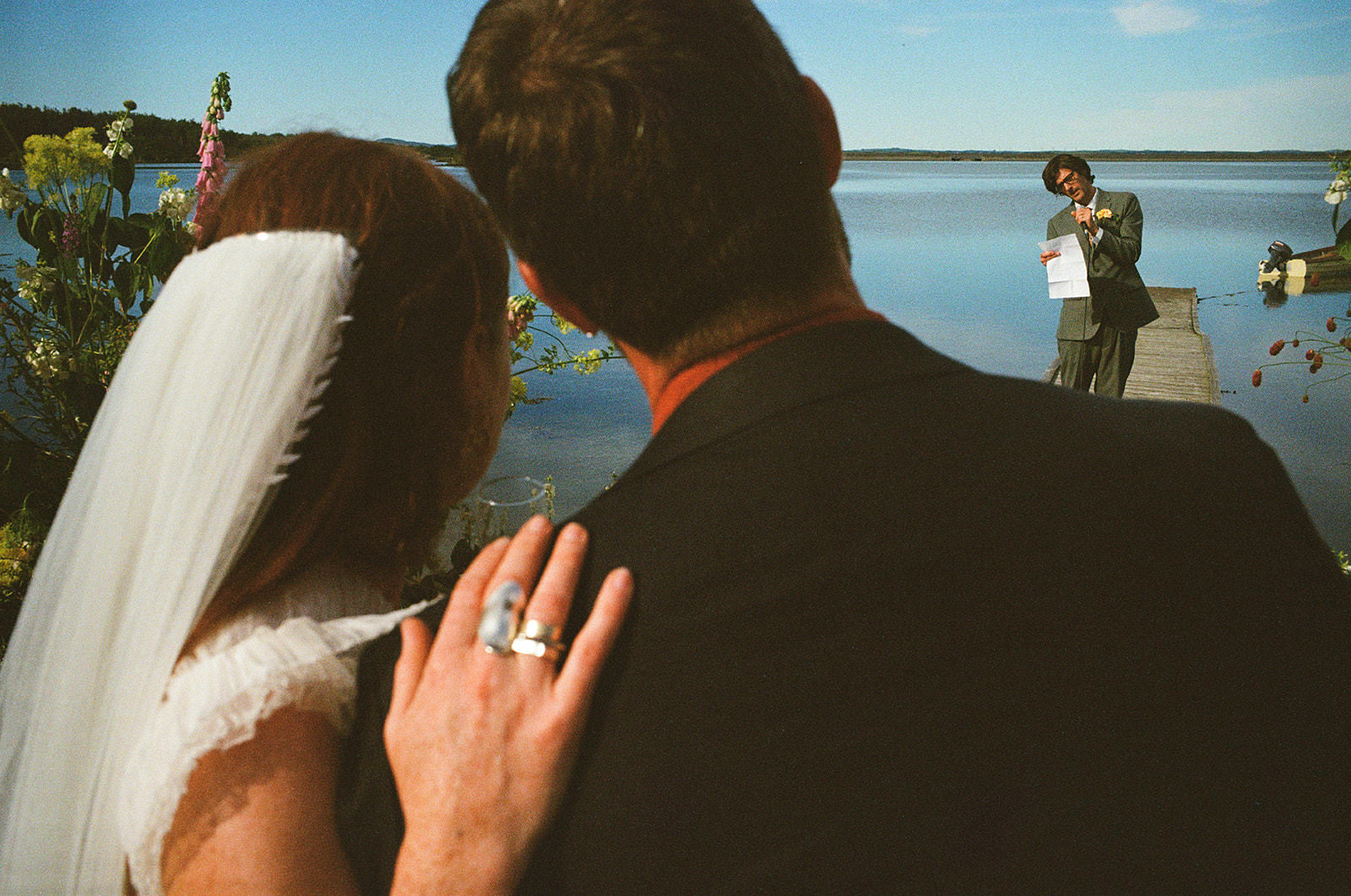 Bride rests hand on groom’s shoulder as vows are read on a quiet lakeside dock.