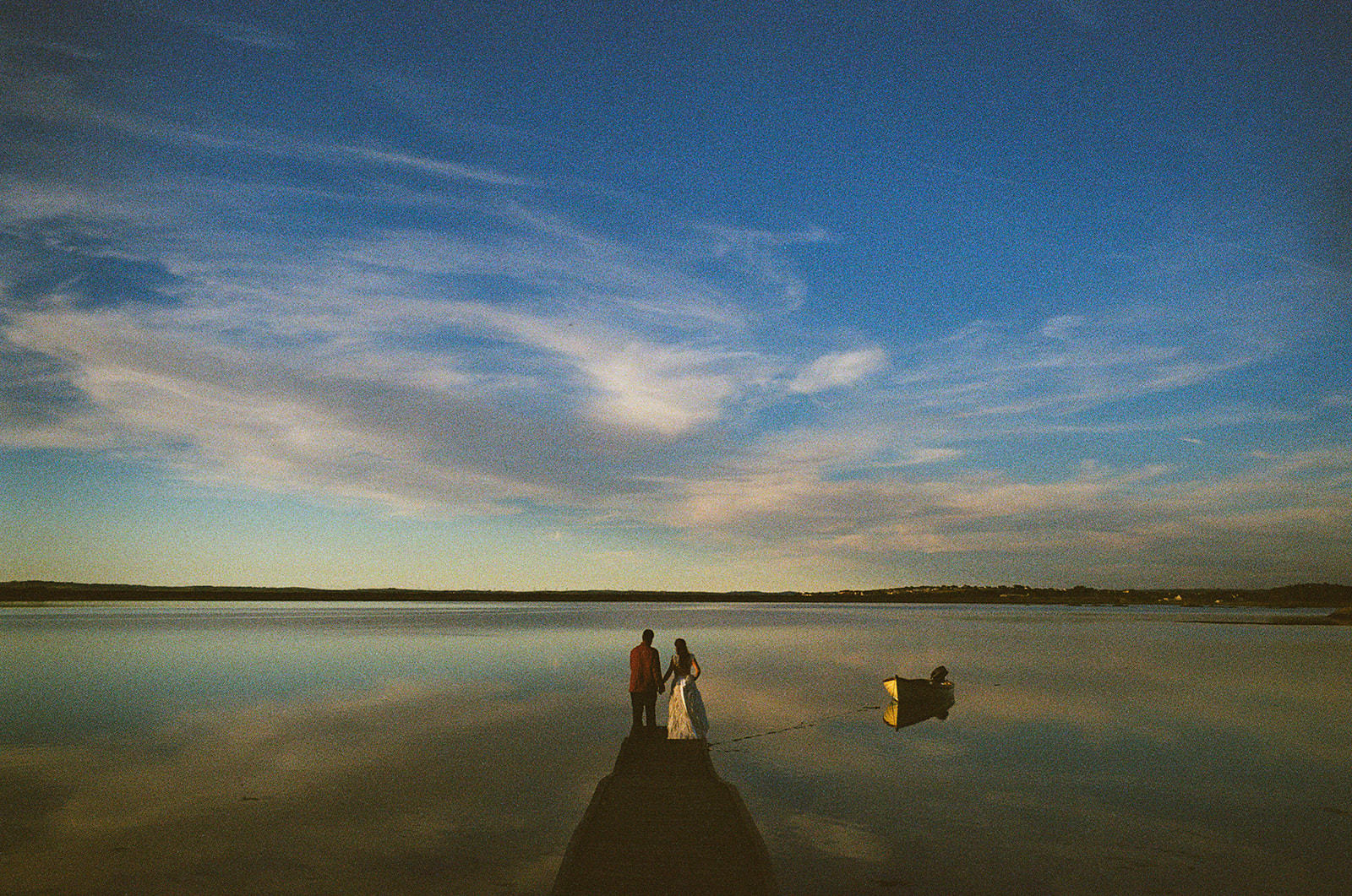 Bride and groom holding hands on a quiet jetty at dusk, reflected in still water.