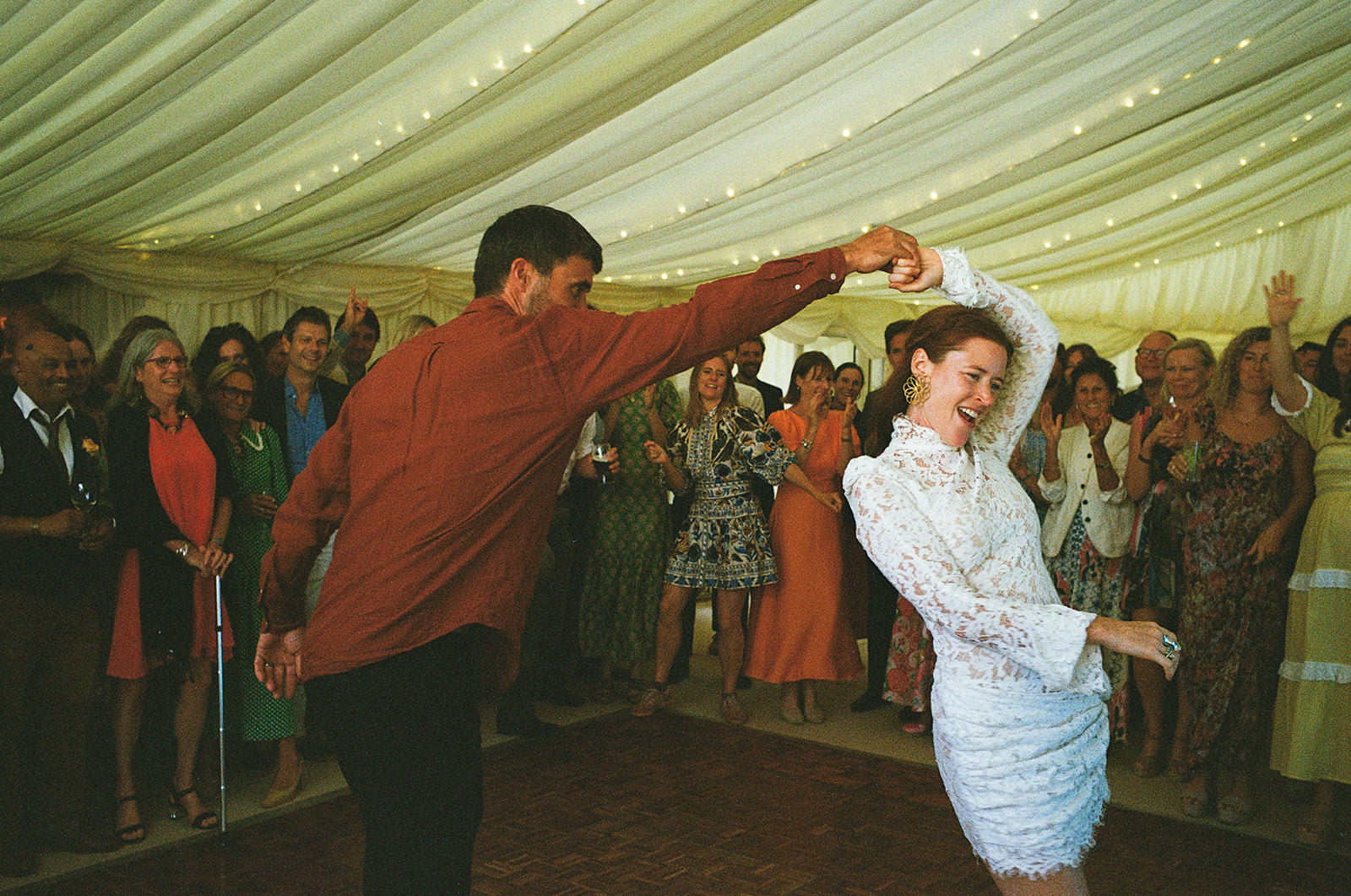 Bride twirls on dancefloor as guests cheer beneath softly lit marquee at wedding reception.