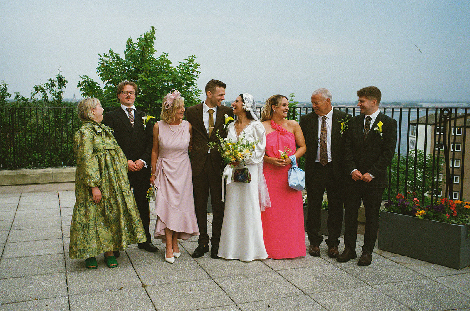 Bride and groom laughing with family on terrace, bouquets in hand, summer wedding day.