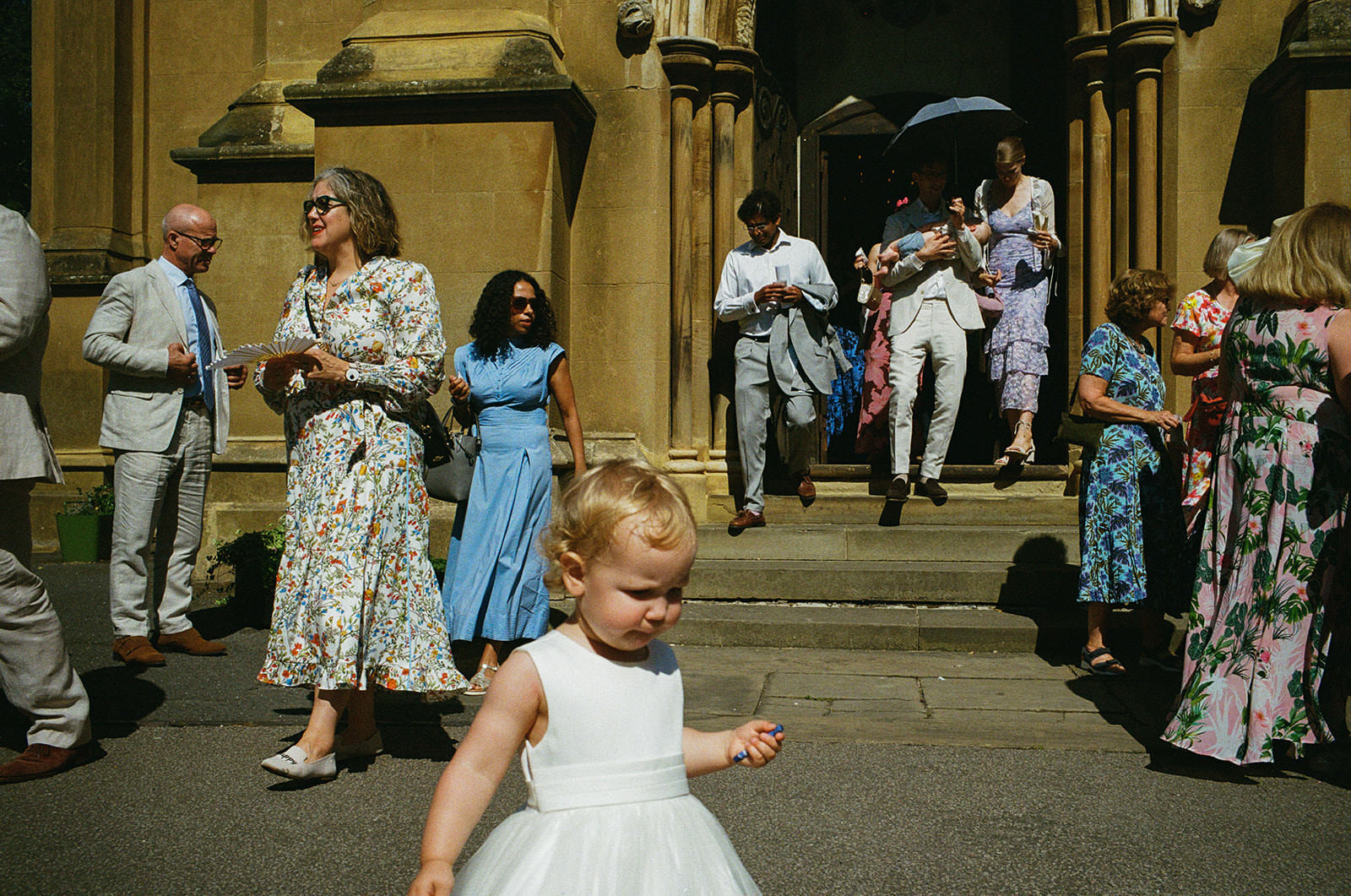 Toddler in white dress wanders past guests leaving church in bright afternoon sun, documentary wedding photography.