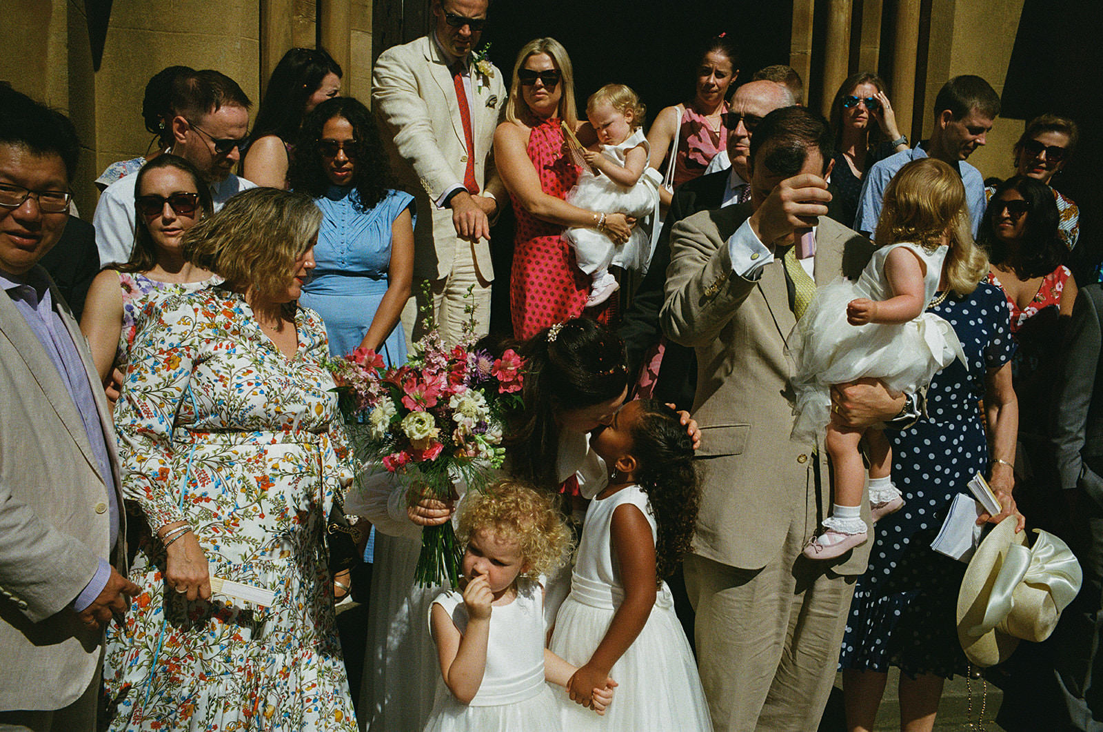 Bride holding bouquet kisses young girl as guests gather in bright church doorway, documentary wedding photography.
