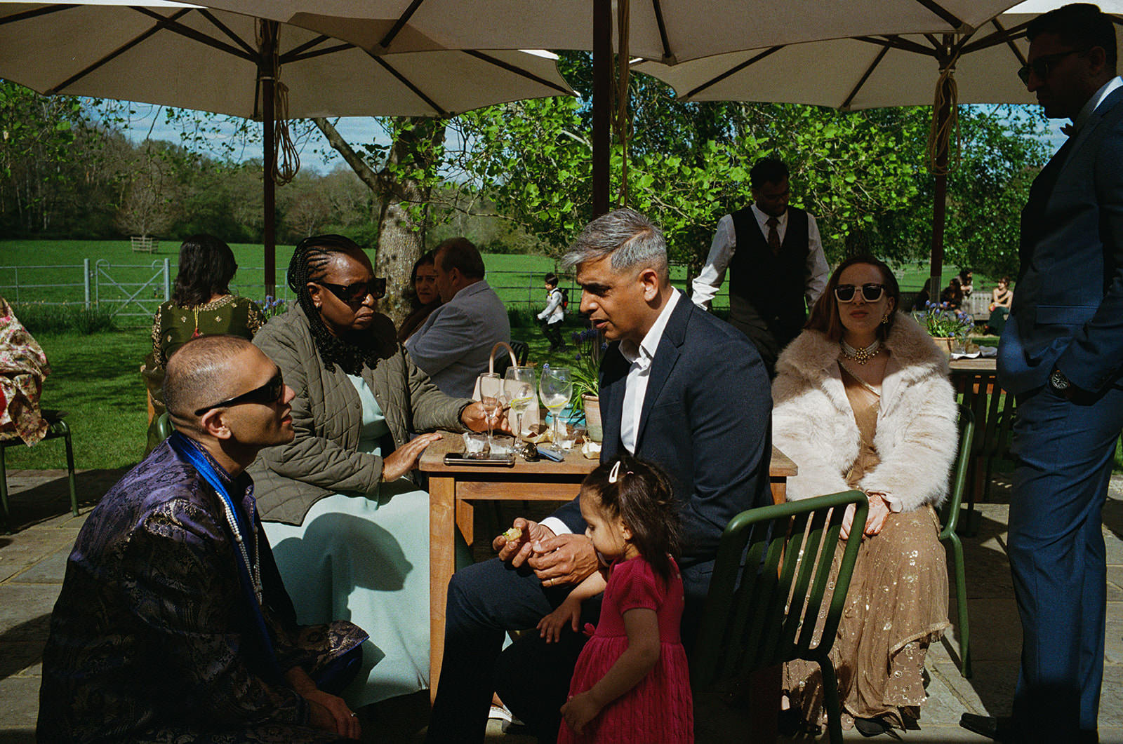 Guests chat at garden wedding reception as a child in red dress stands beside the table.