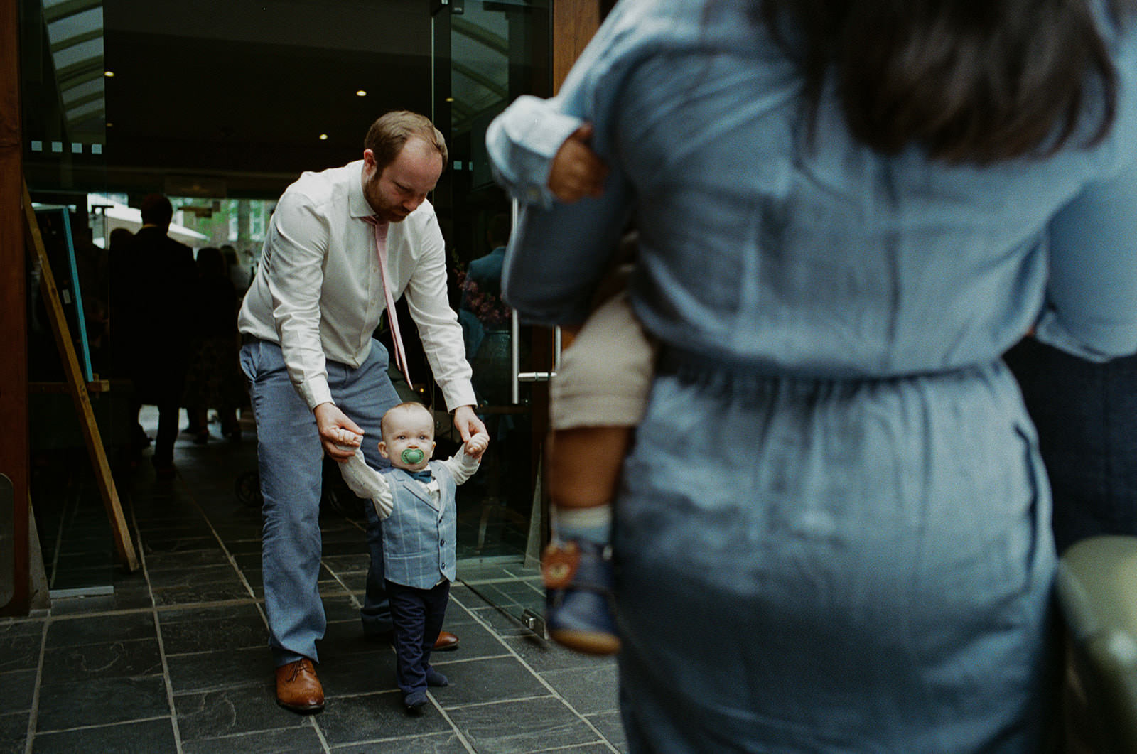 Father steadies toddler in waistcoat outside wedding venue, small steps across stone floor.
