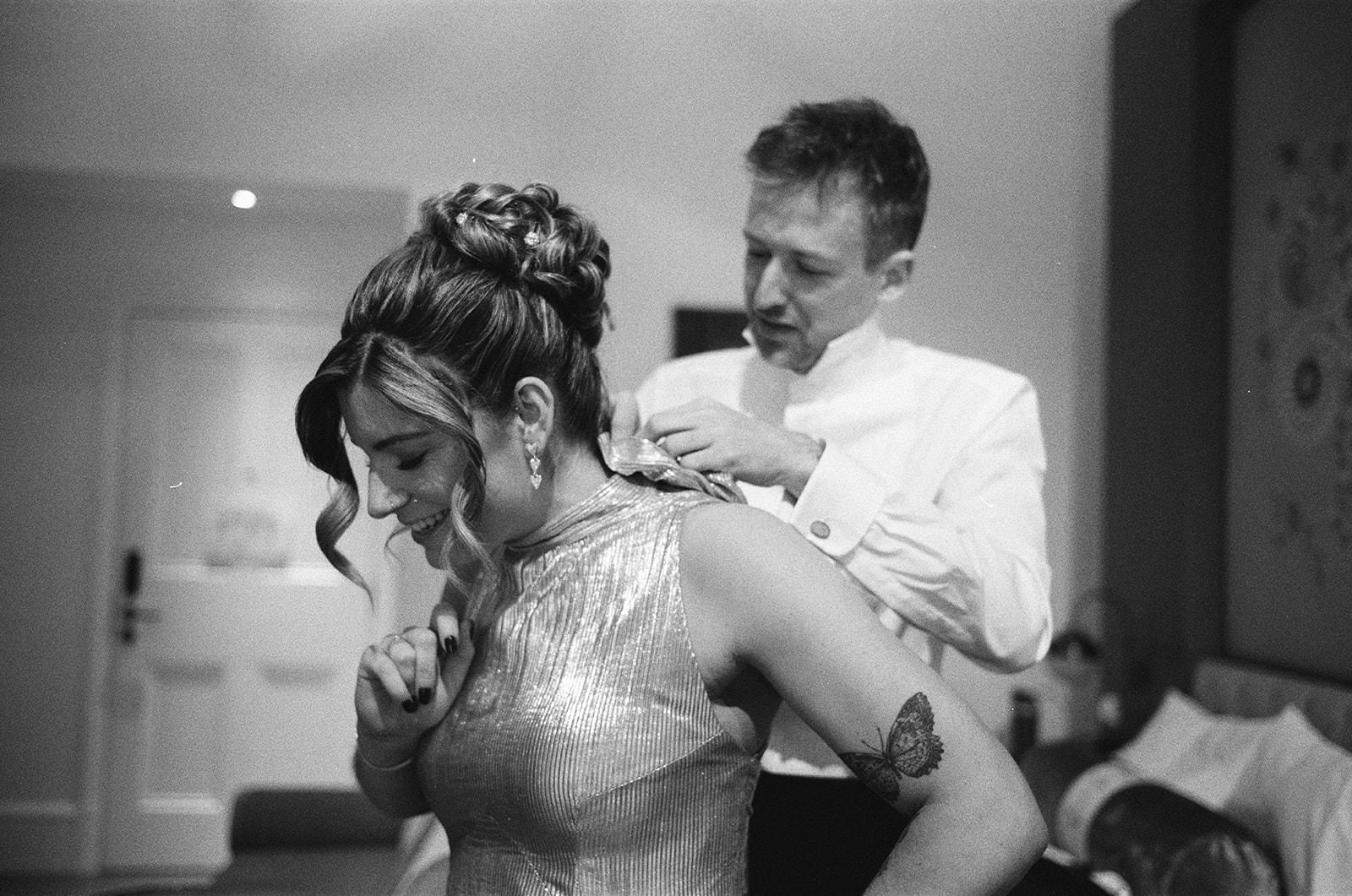 Bride smiling as partner fastens her dress in quiet room, documentary wedding photography moment.