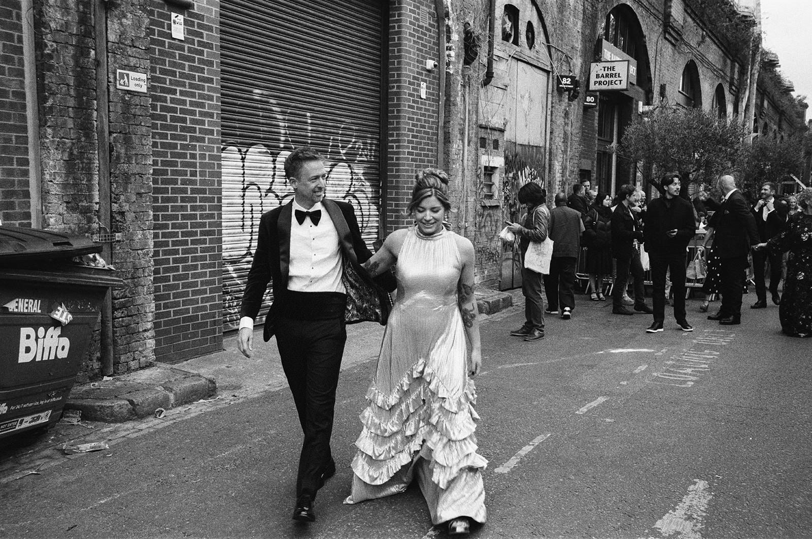 Bride and groom walk hand in hand along city street, guests gathered outside brick venue, documentary wedding photography.