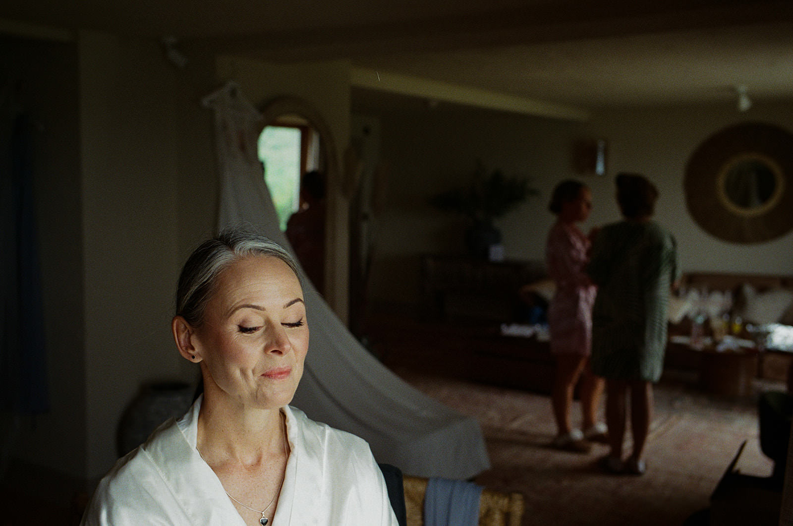 Bride sits quietly with eyes closed as veil falls behind, friends chatting softly in background.