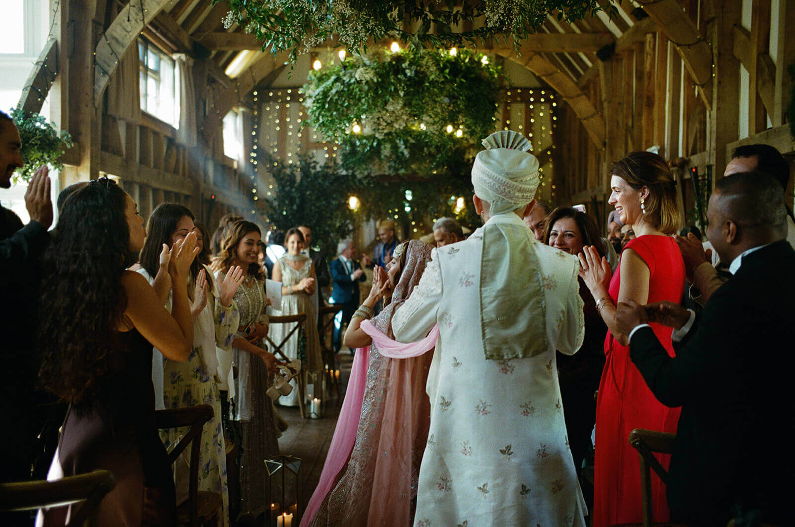 Guests clap as couple walk beneath greenery in candlelit barn wedding ceremony.