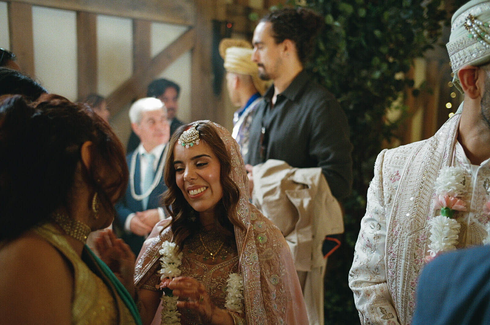 Bride smiling during lively Indian wedding ceremony, floral garland in hand, guests gathered close.