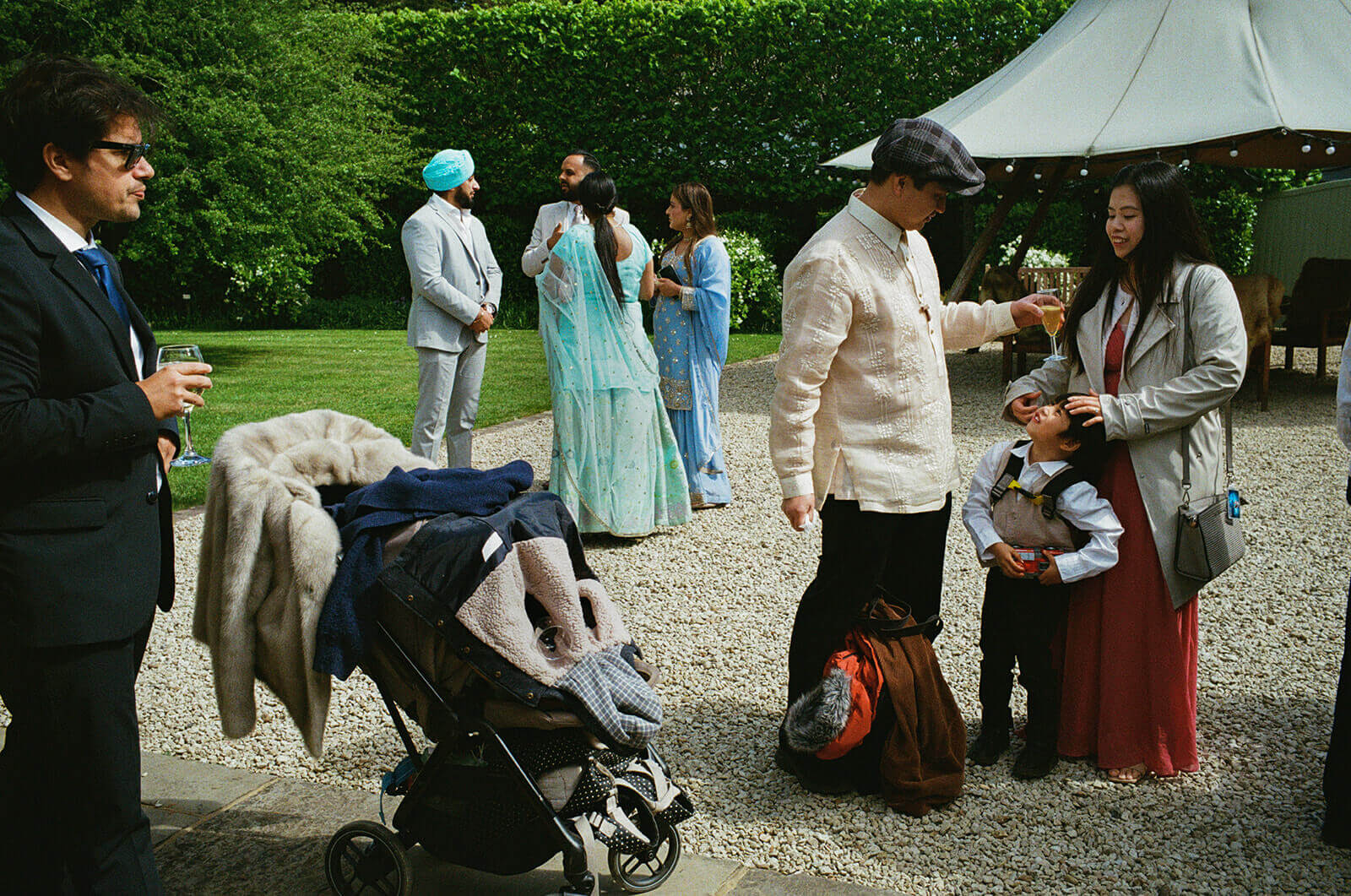 Guests chatting on a gravel lawn as a child looks up, sunlight catching a quiet wedding moment.