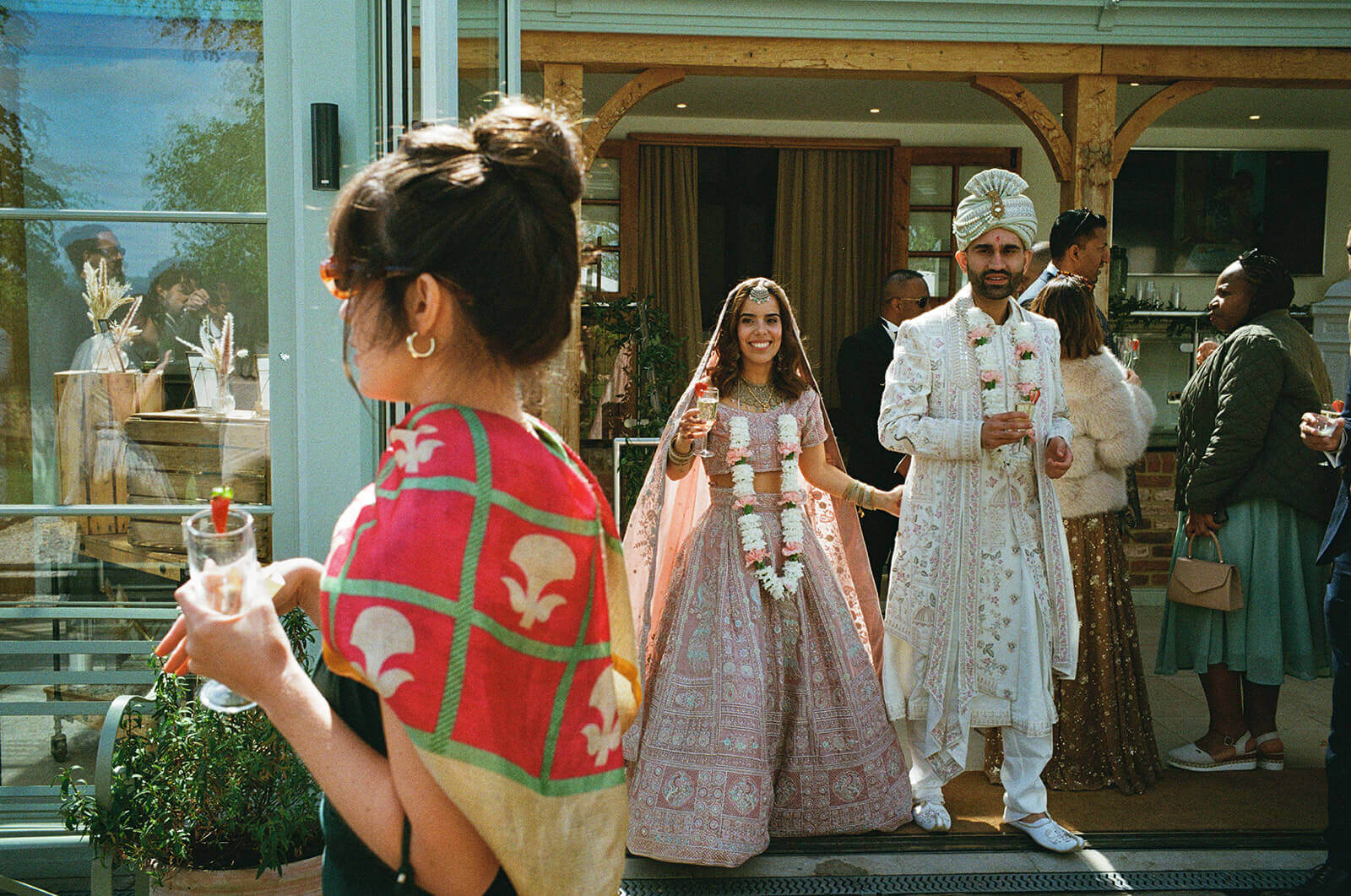 Indian wedding couple step outside, smiling with drinks as guests gather behind.