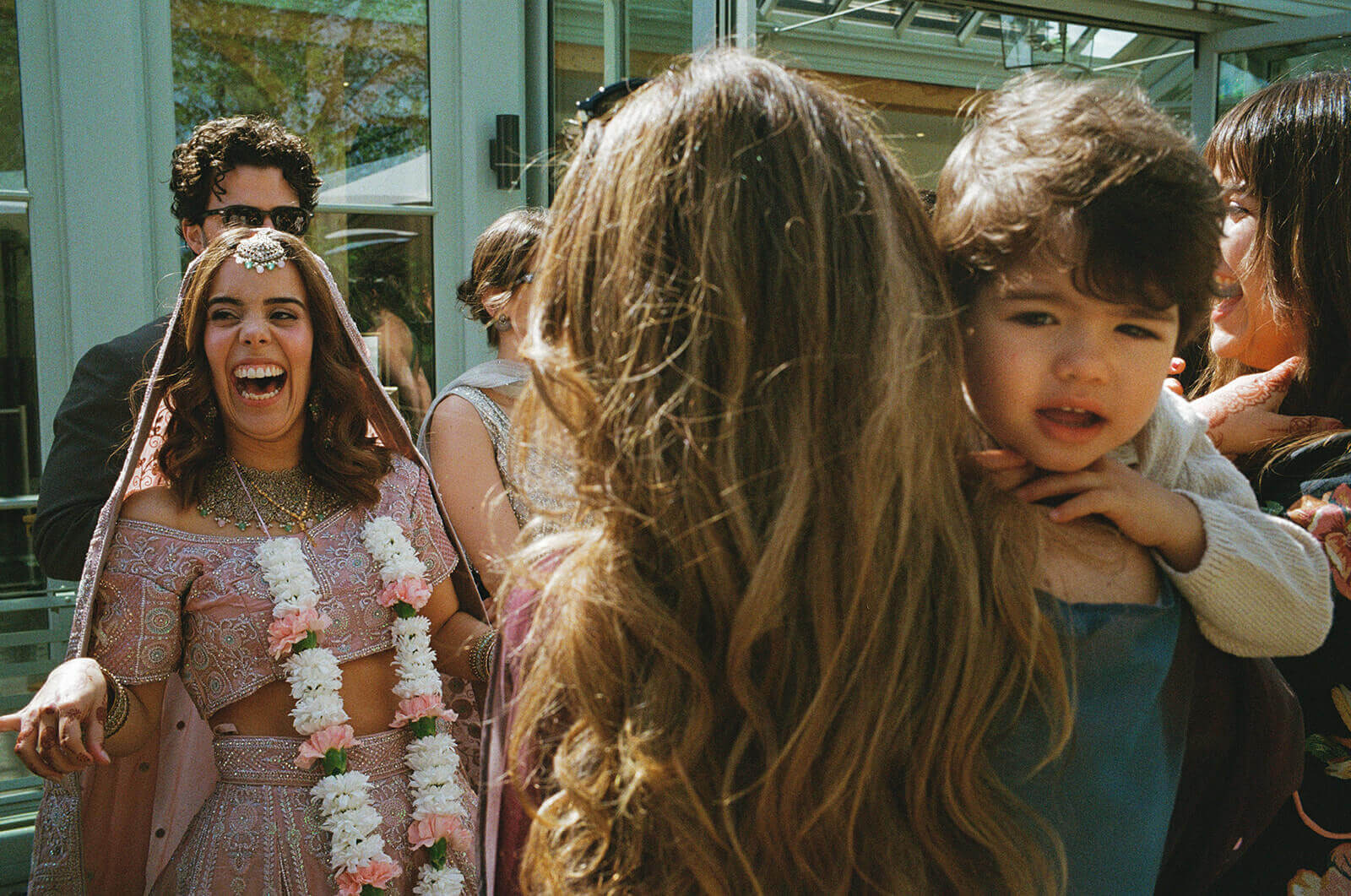 Bride laughing in pink sari as family embrace in sunlit courtyard, documentary wedding photography.