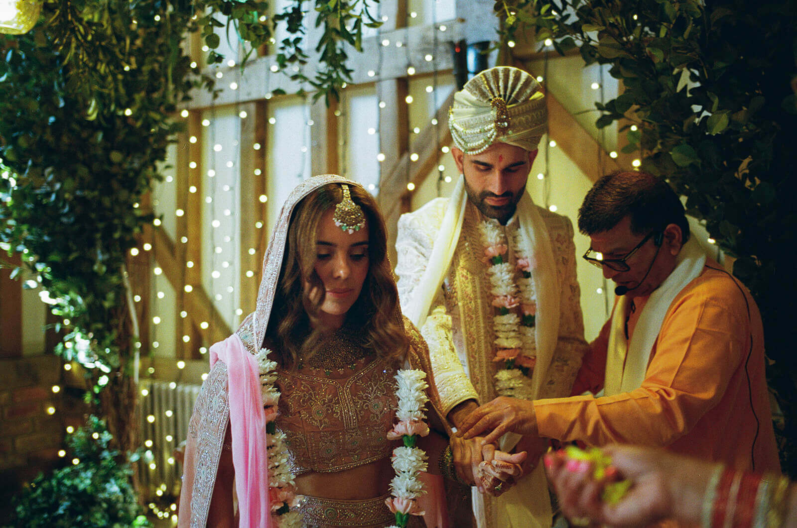 Hindu wedding ceremony beneath fairy lights as couple exchange garlands, priest guiding their hands.