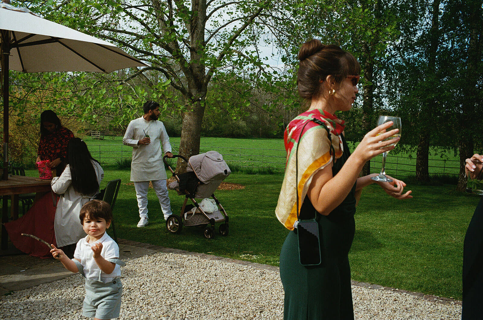 Child clutching a stick wanders across garden lawn as guests chat and sip drinks at wedding reception.