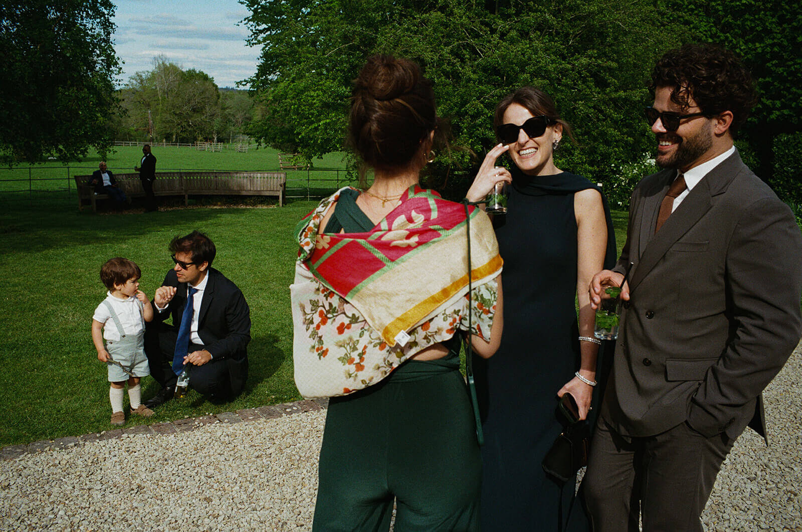 Guests laugh in sunny garden, drinks in hand, during relaxed documentary wedding photography moment.