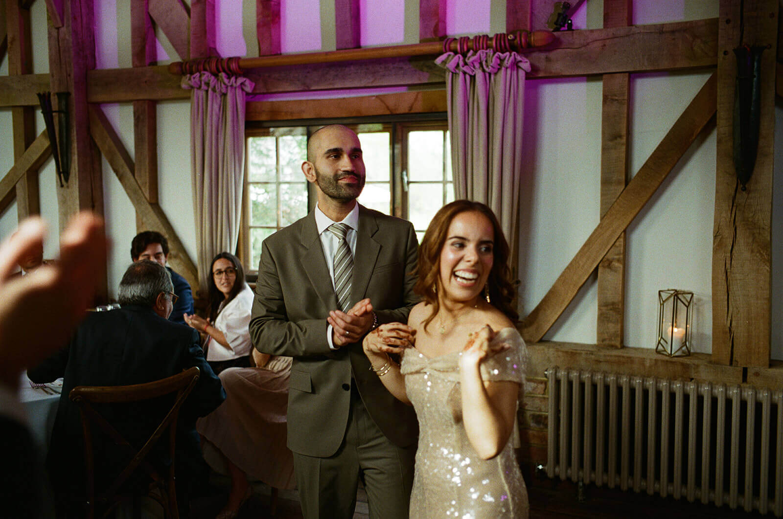 Bride laughing as groom claps during wedding reception in timber-framed hall.