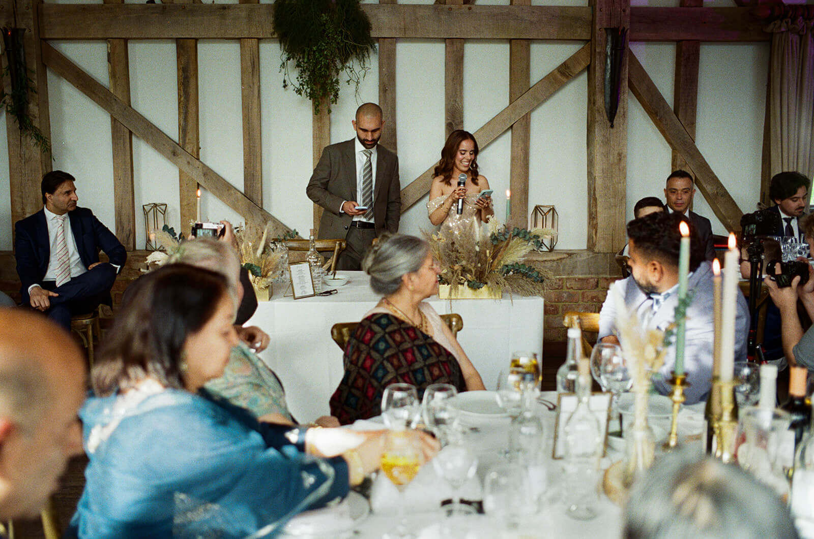 Bride reads a speech, smiling, as guests listen at candlelit barn wedding reception, captured in documentary wedding photography.