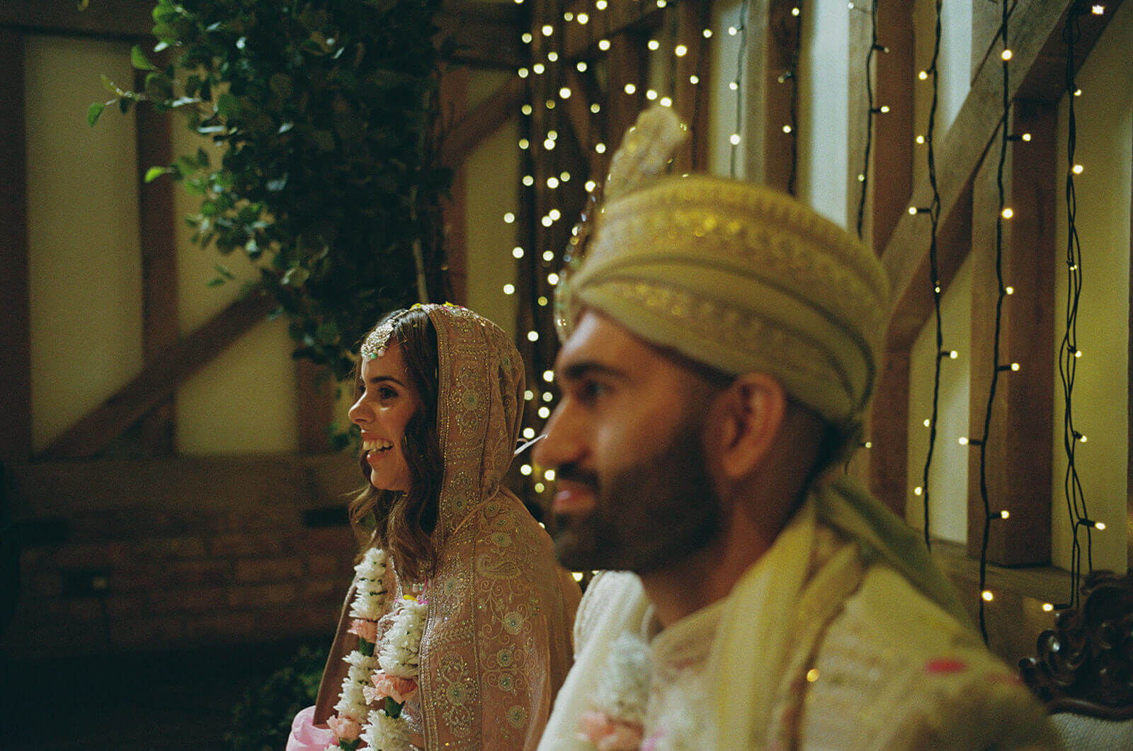 Bride and groom smiling beneath warm fairy lights during evening wedding ceremony.