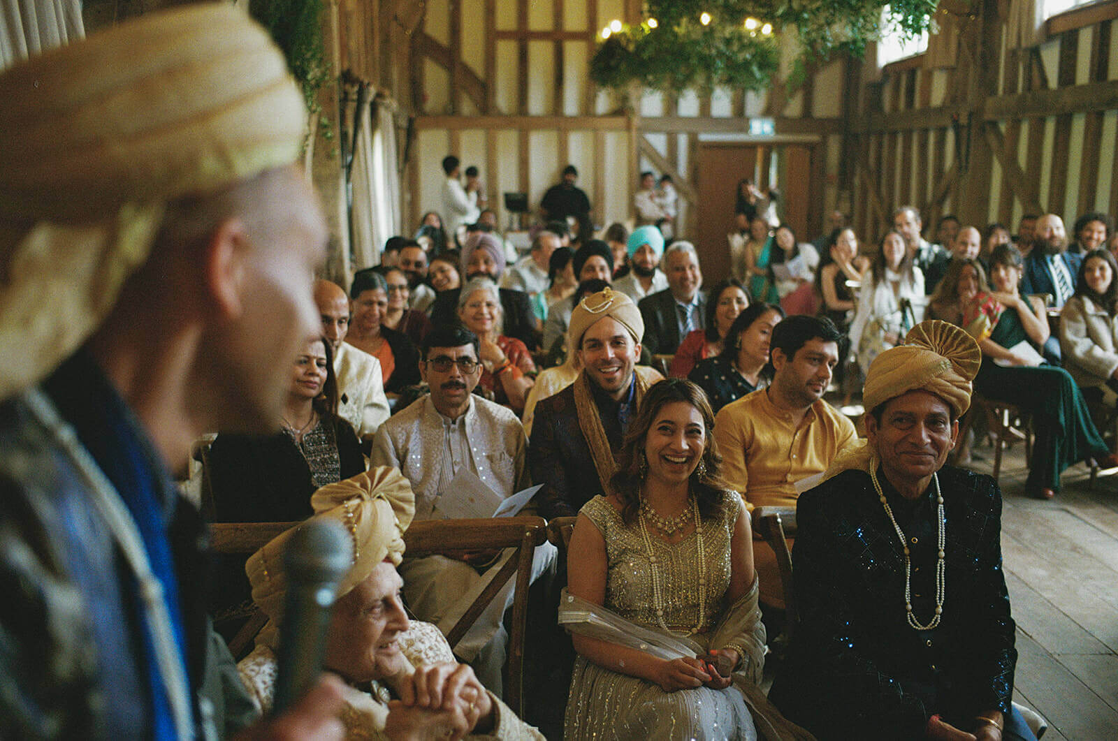 Guests smiling during wedding ceremony inside timber hall as groom speaks in foreground.