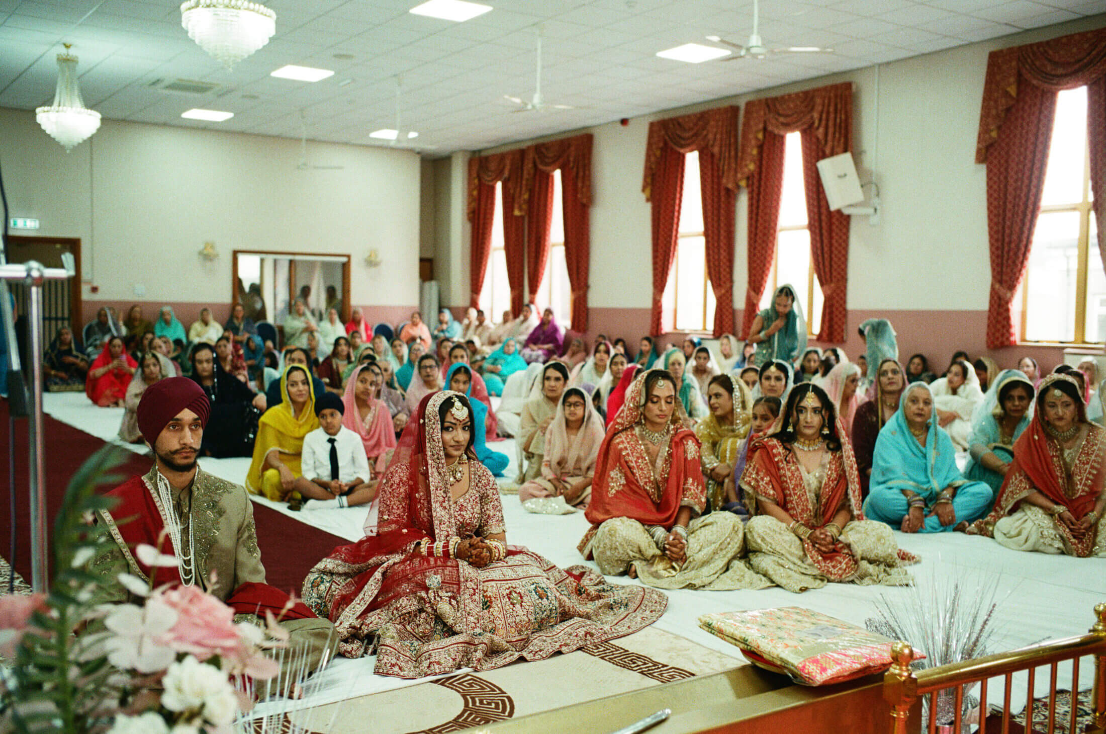 Sikh bride and groom seated during their wedding ceremony, surrounded by guests. Captured on 35mm film