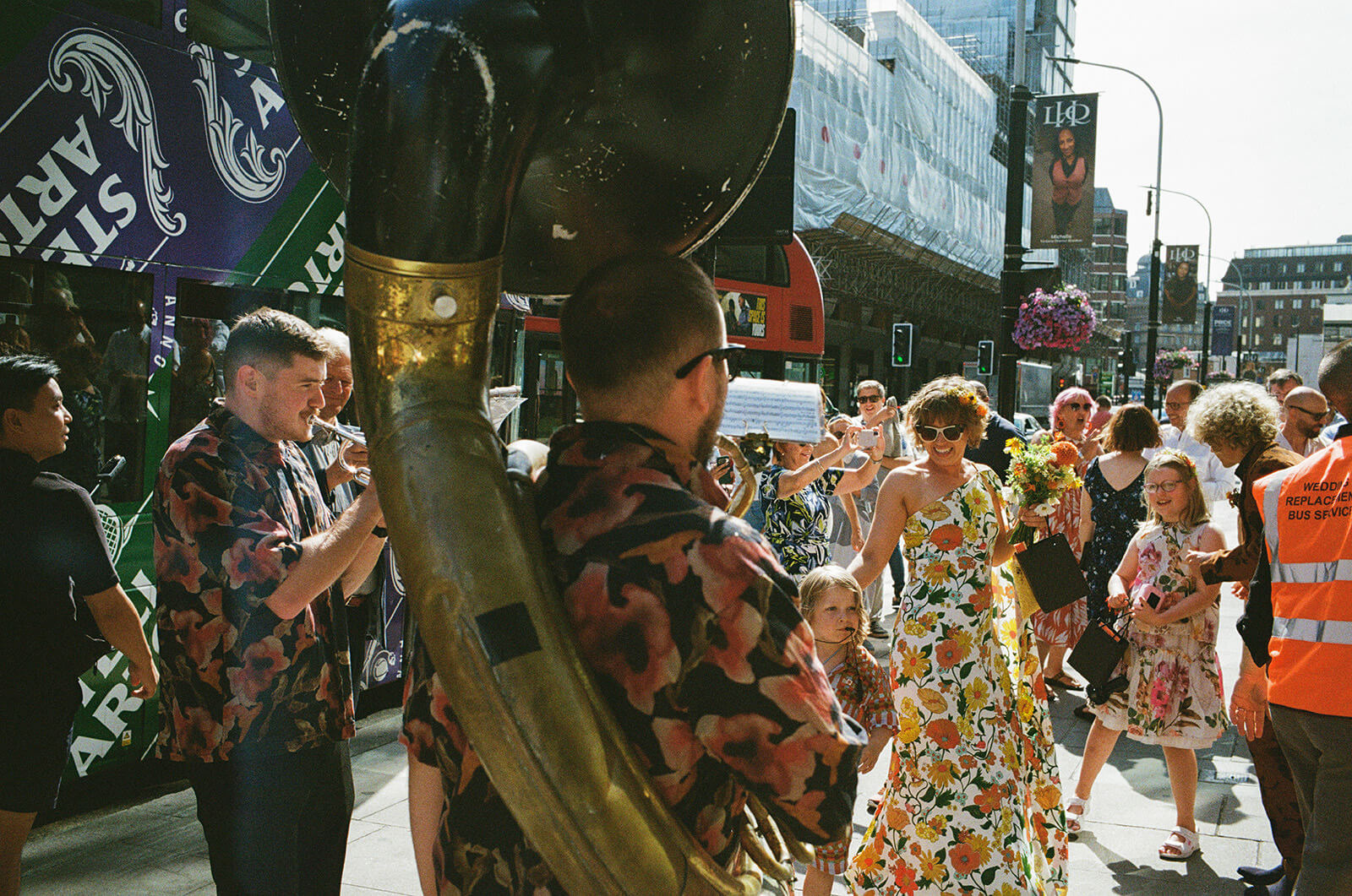 Wedding band greets the guests as they make their way through the streets to a London wedding venue. In the centre the bride wears a non-traditional, colourful floral dress, and in the foreground we see a Sousaphone. Captured on 35mm Analogue Film