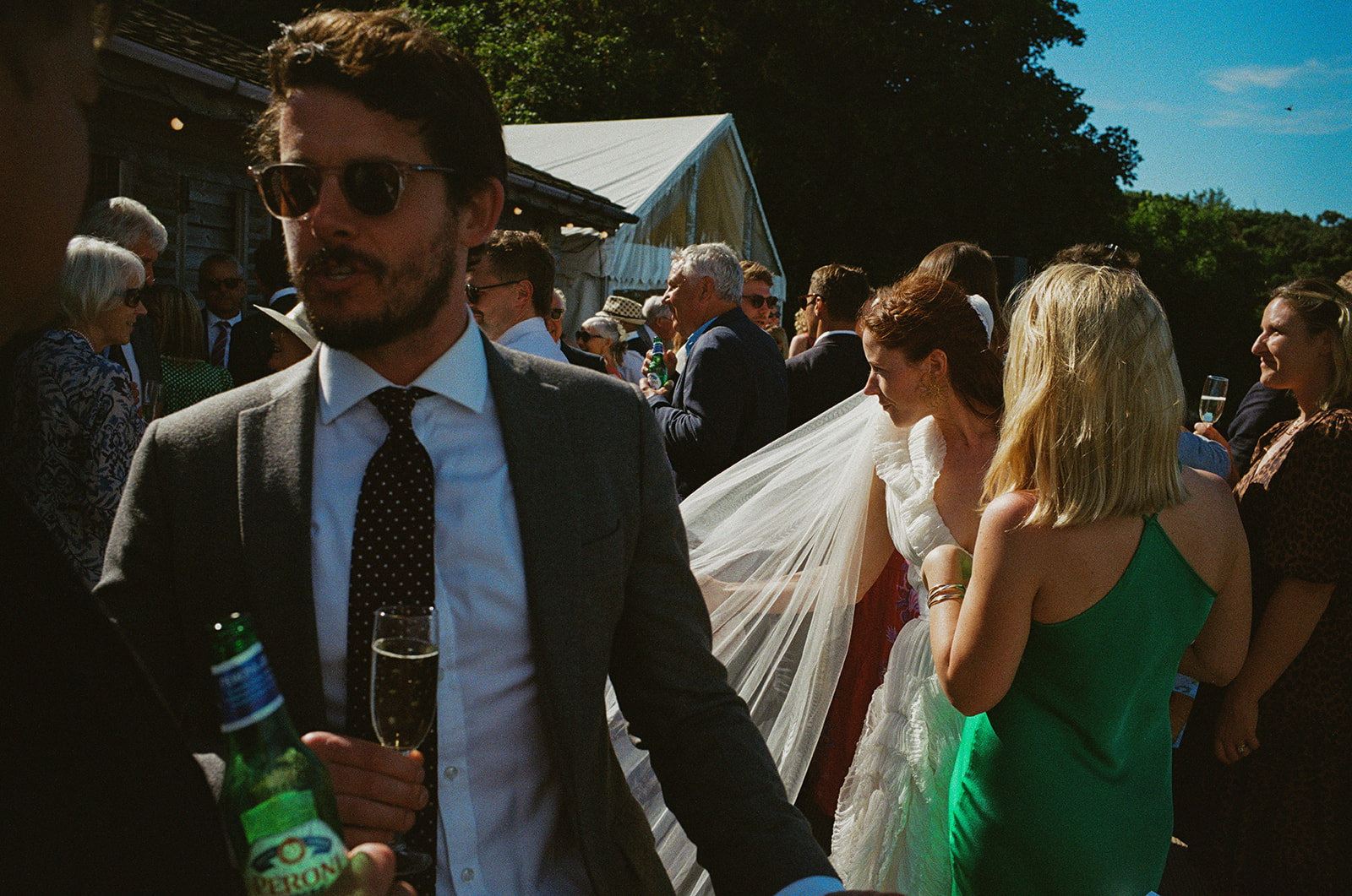 Guests mingle in summer sunshine as bride's veil drifts through outdoor wedding reception, captured in documentary wedding photography.