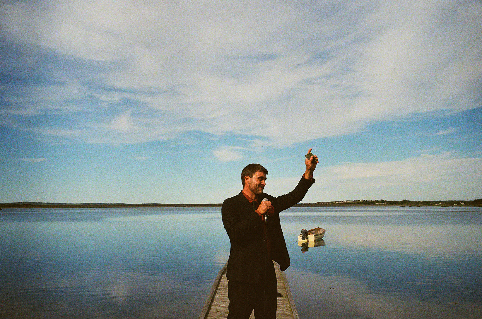 Man giving a wedding speech on a lakeside jetty, raising a glass beneath wide evening skies.