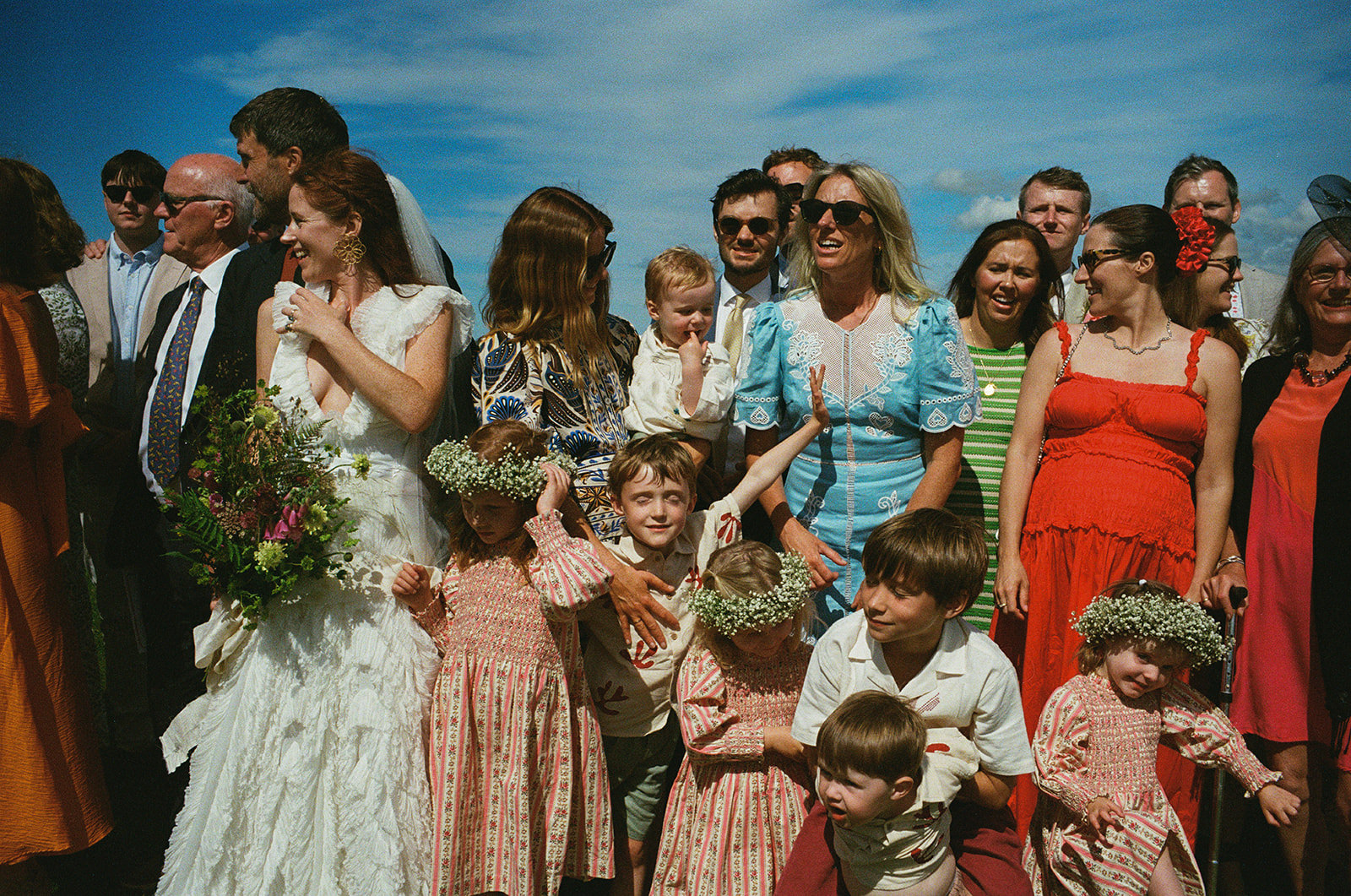 Documentary wedding photography captures bride and guests laughing as children in flower crowns gather under blue summer sky.