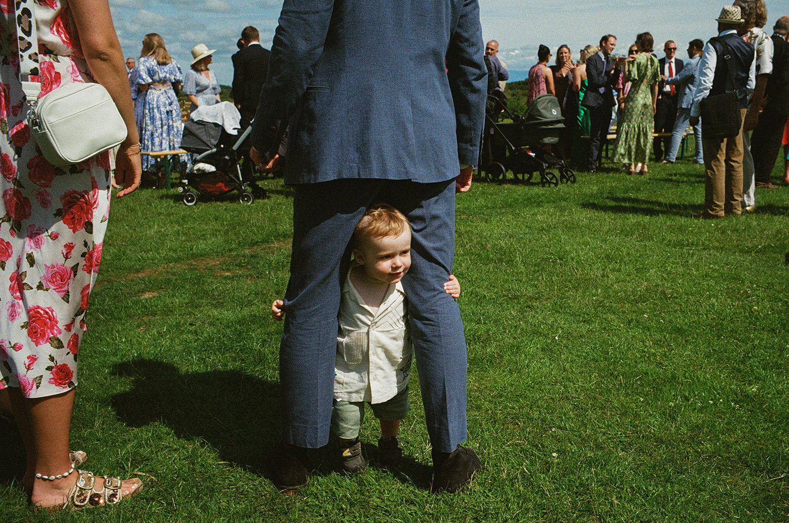 Toddler peeks between man’s legs on sunny lawn as wedding guests mingle nearby.
