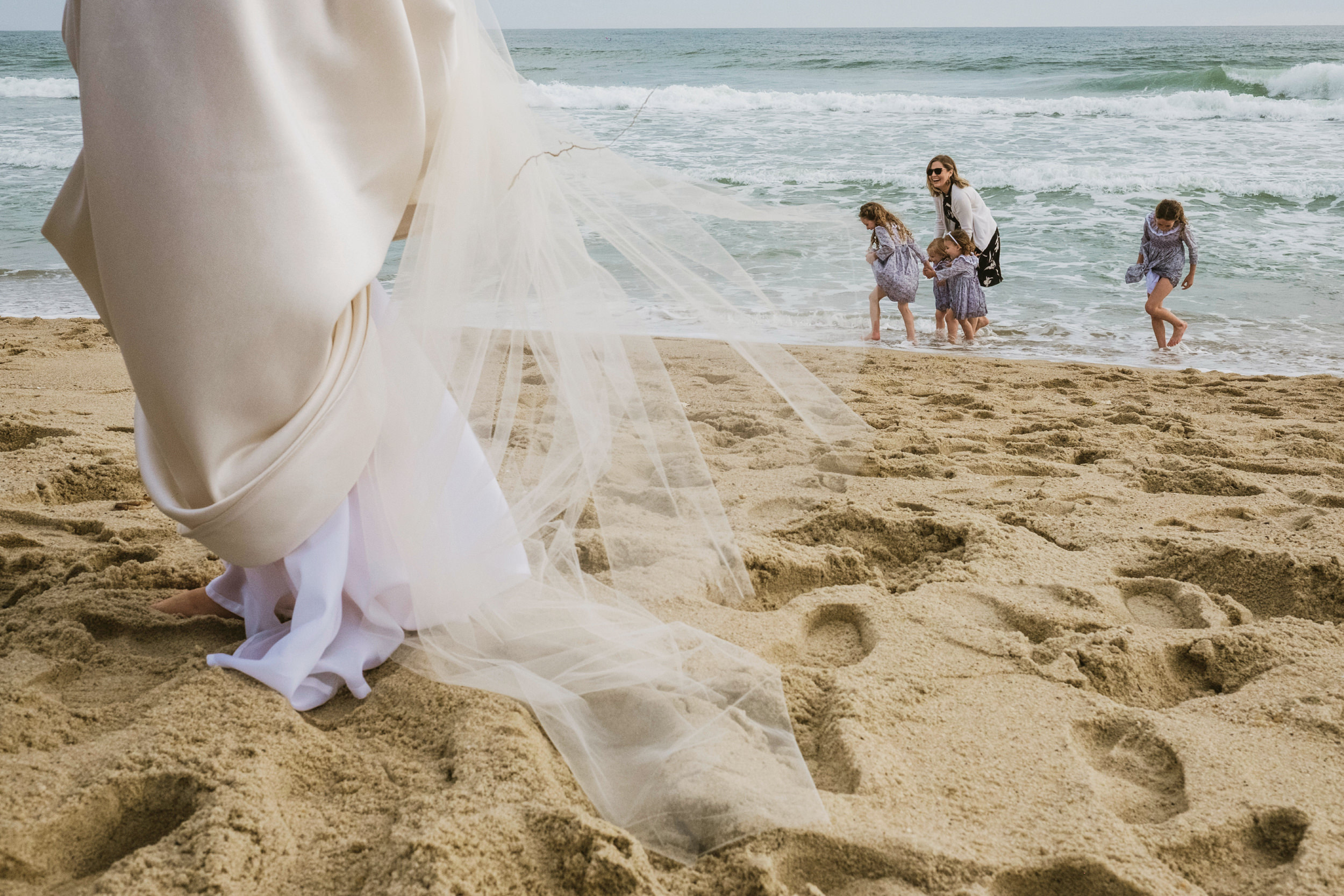 A close-up shot of a wedding dress and veil dragging through the deep sand on a beach, with flower girls playing at the shoreline in the blurred background.