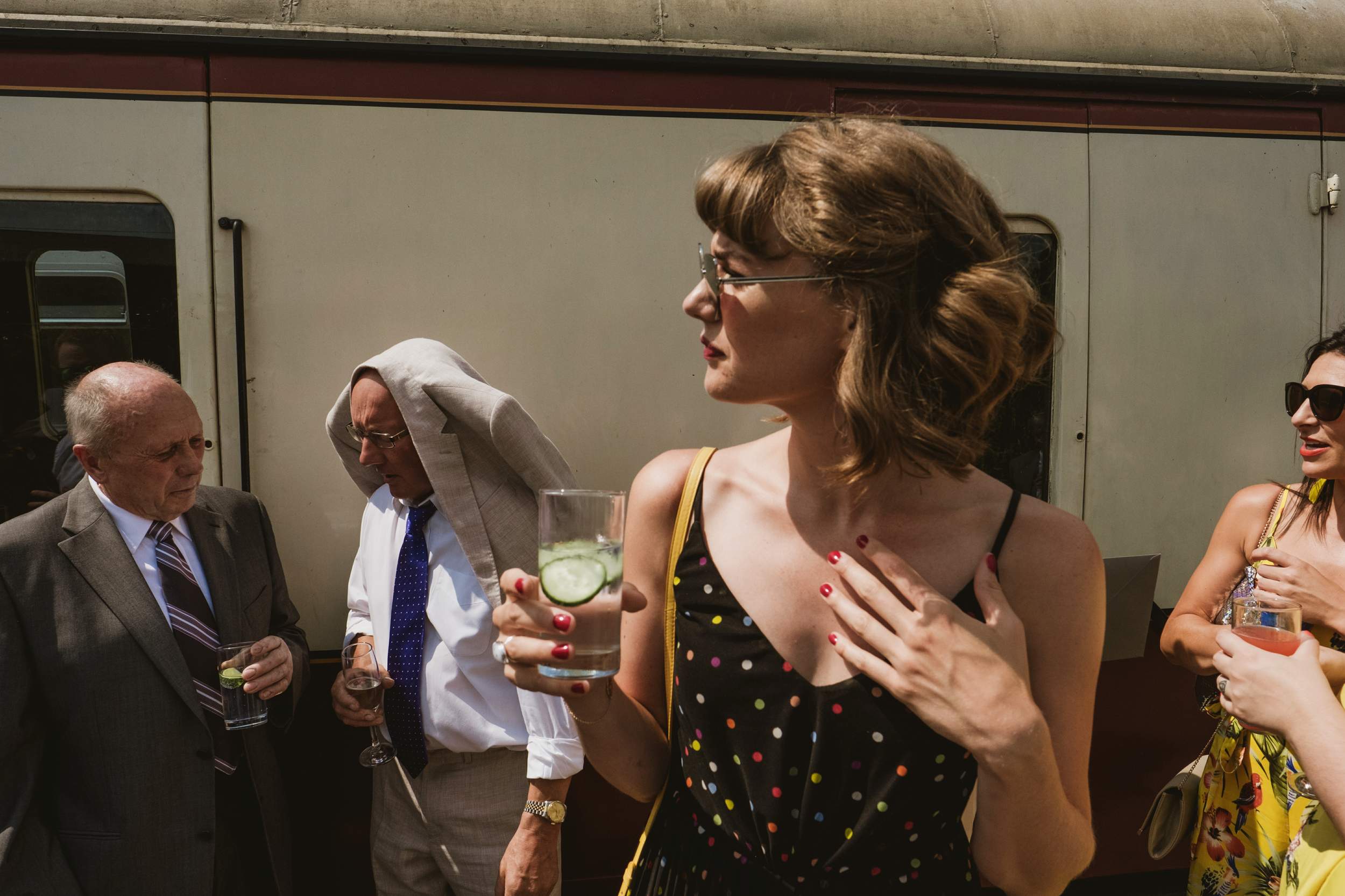 A candid, documentary-style photograph of wedding guests standing beside a vintage cream and maroon train carriage in bright, direct sunlight. In the foreground, a woman in a black polka-dot dress holds a glass with cucumber slices, her face in profile as she looks away. Behind her, a man wears his light-coloured suit jacket over his head to shield himself from the sun, while other guests talk and hold drinks in the sharp shadows cast by the train.