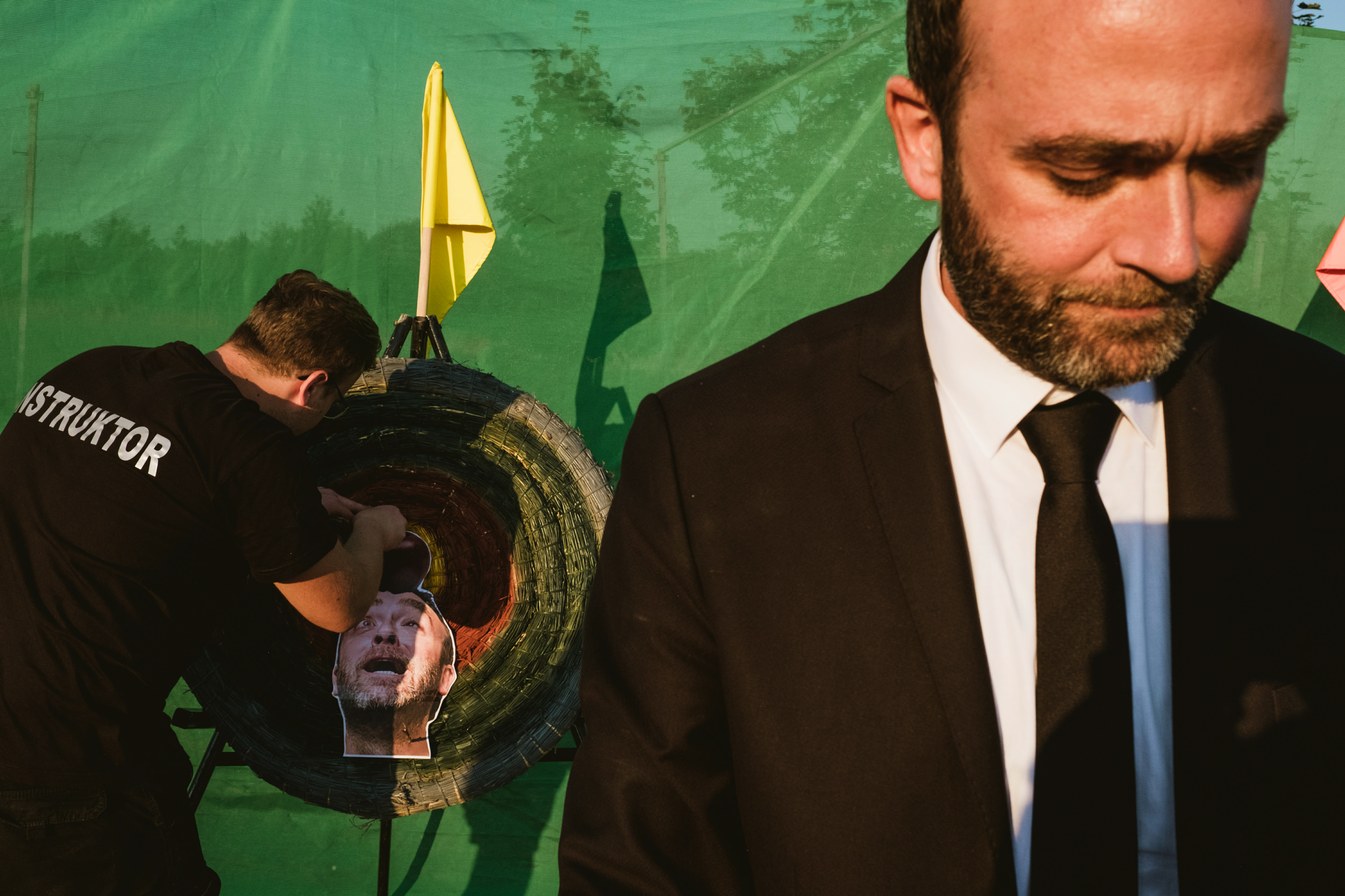 A documentary-style photograph at a wedding in Poland showing a groom in a black suit and tie standing next to an archery target. An instructor in a black t-shirt is pinning a cardboard cutout of the groom's face onto the bullseye. The scene is set against a green mesh backdrop with a yellow flag visible.