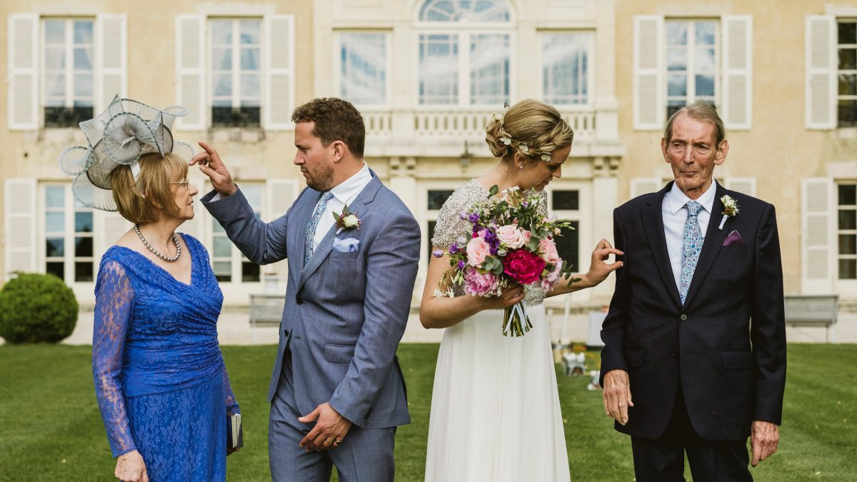 Unposed, documentary interpretation of a group photograph - the bride and groom stand in the centre looking outwards towards their parents. The couple tweak the outfits of the parents ready for a more formal group shot.