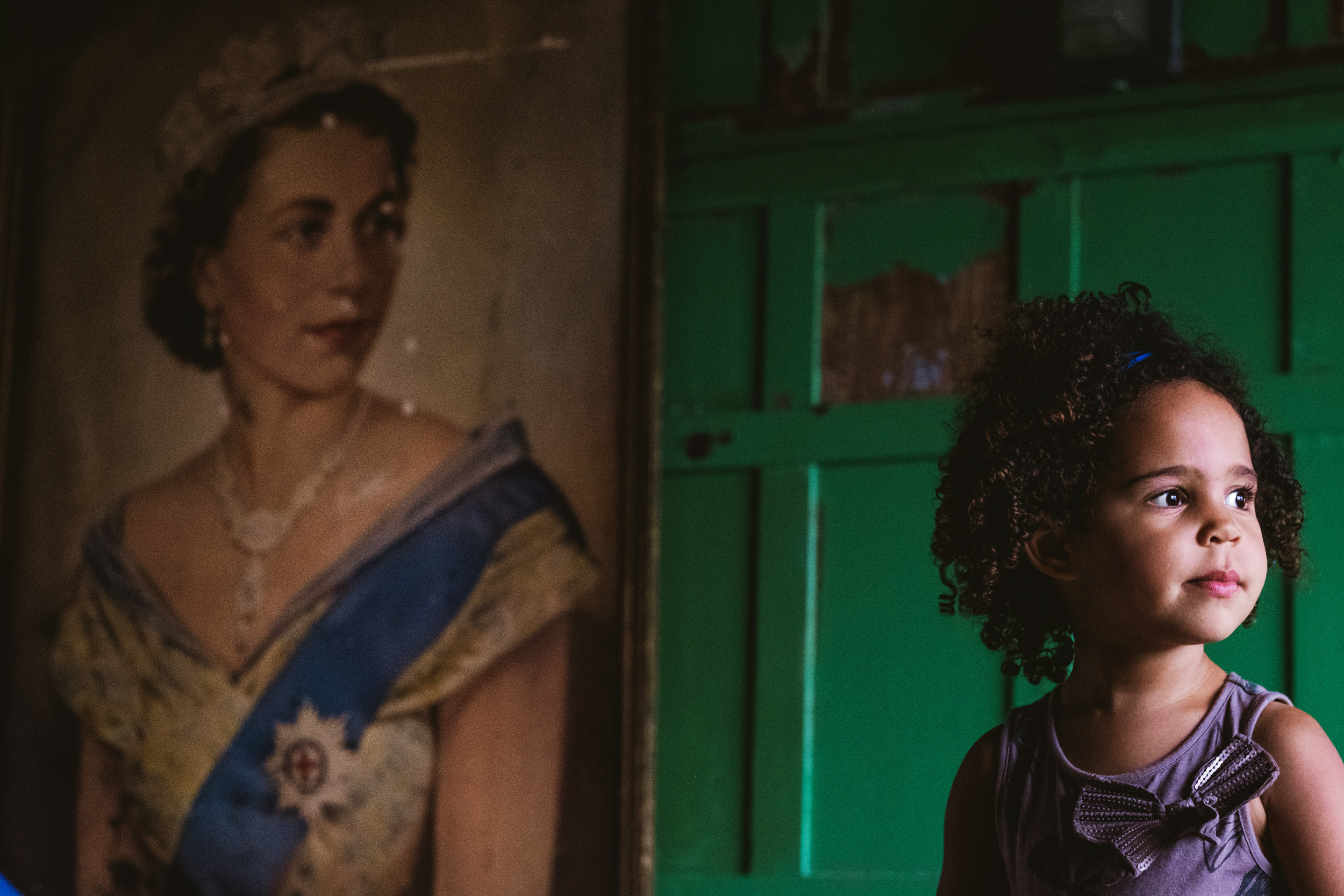 A tight, profile shot of a young girl with dark, curly hair and a small blue hair clip, looking intently to the right of the frame. She is positioned against a vibrant, rustic green wooden door with peeling paint. In the soft-focused background on the left, a large, vintage oil painting portrait of a young Queen Elizabeth II hangs on the wall, gazing in the opposite direction.