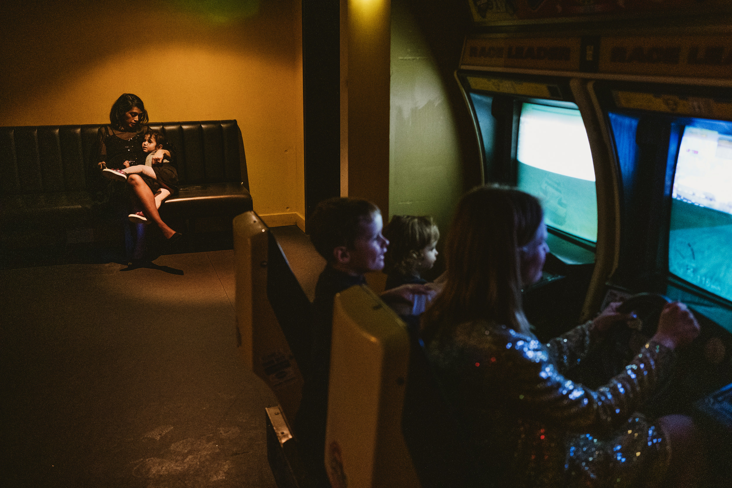 A cinematic documentary wedding photograph at Bloomsbury Lanes, London. The frame is split by light and mood: on the right, the scene is bathed in a cool, electric blue neon glow from arcade racing games. On the left, a mother and child are tucked into a dark leather booth, illuminated by a warm, golden spotlight as they share a quiet hug. The image highlights the visceral contrast between the frantic energy of the arcade and a moment of still, human sanctuary.