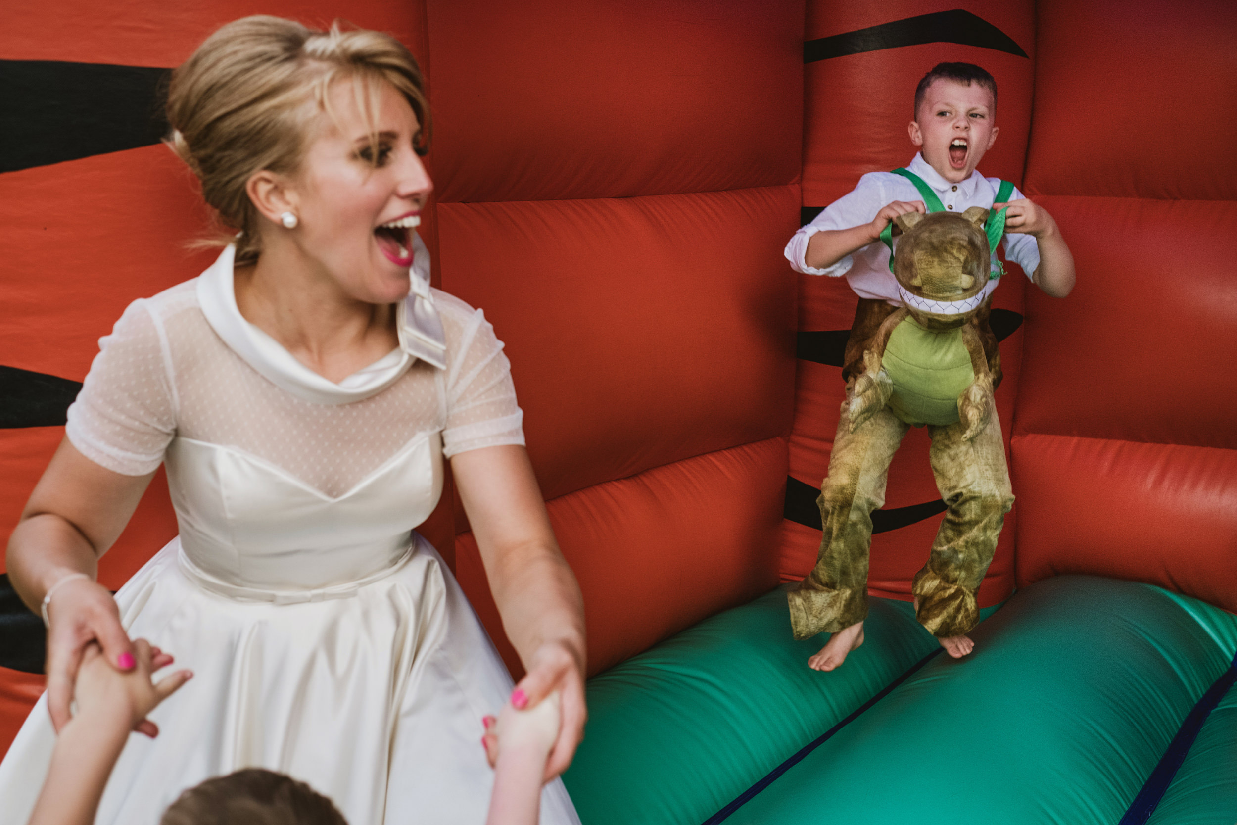 Documentary wedding photography of a bride laughing in a bouncy castle while a child in a dinosaur suit jumps in the background.