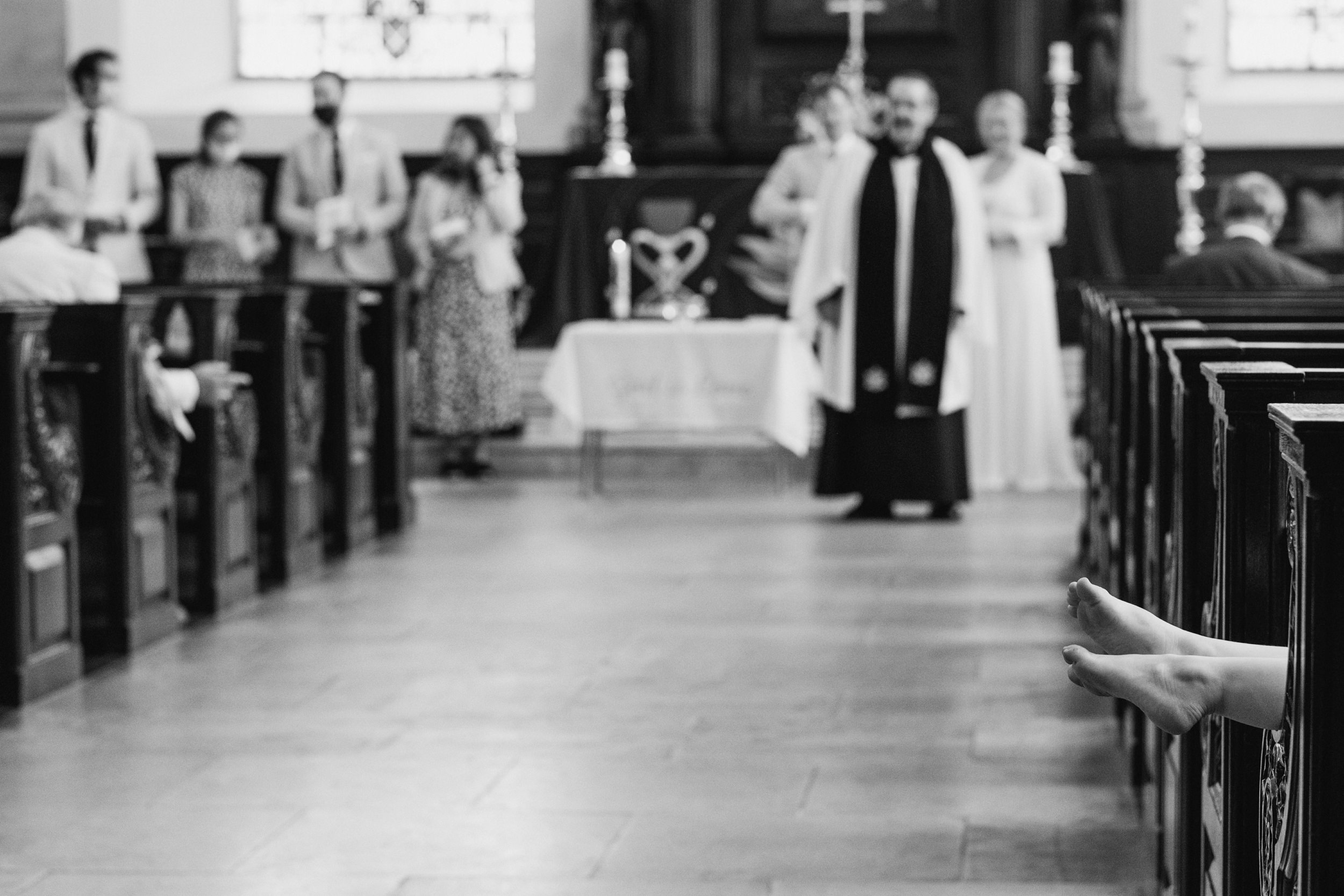 A black and white documentary-style photograph taken inside a church during a wedding ceremony. The background is softly out of focus, showing a vicar in vestments standing near the altar with wedding guests gathered. The sharp focus is entirely on the foreground on the right, where a pair of bare feet extends horizontally out from a dark wooden church pew into the center aisle, capturing a candid and spontaneous moment of comfort during the formal event.