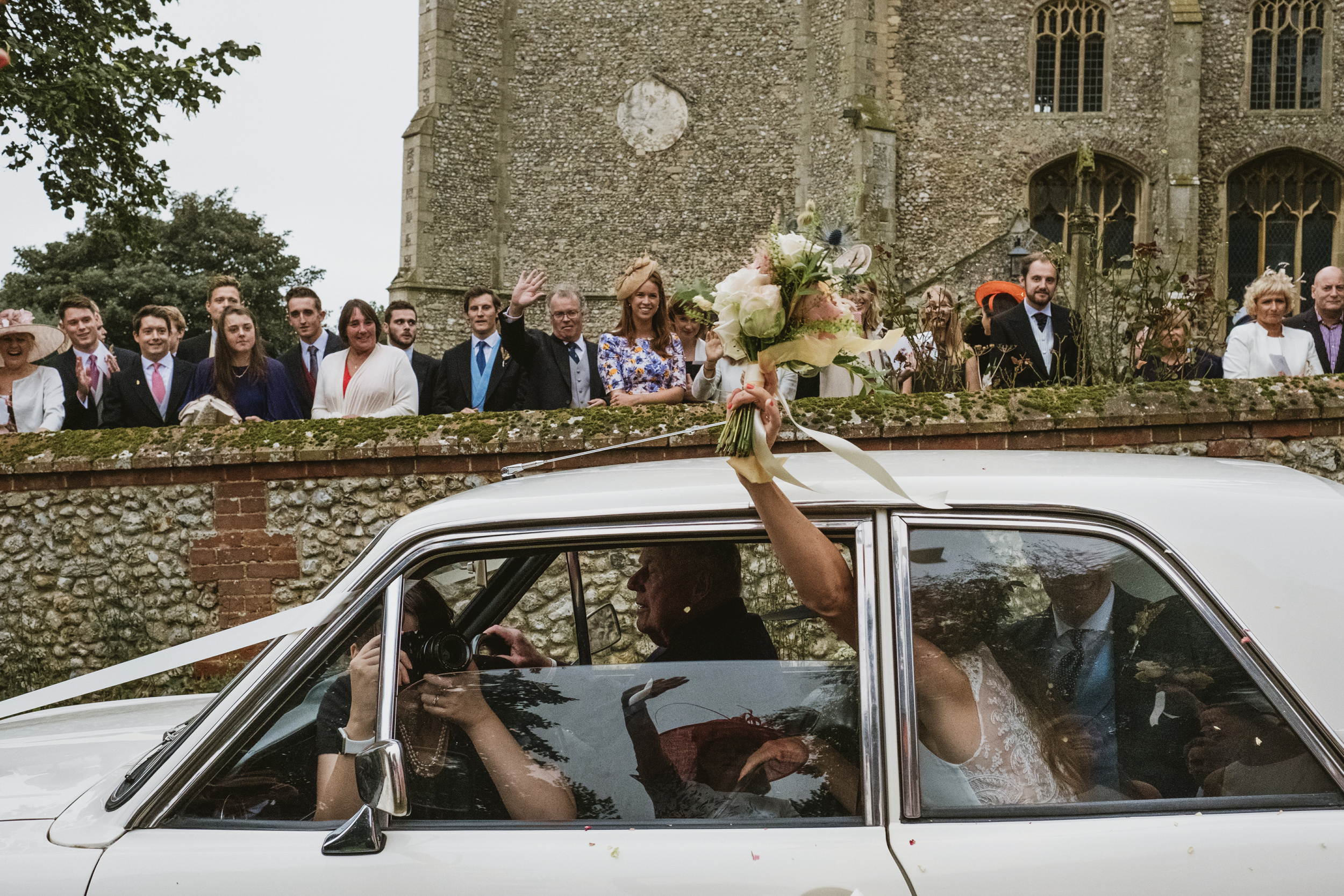 View from inside wedding car showing photographer capturing bride waving bouquet to guests