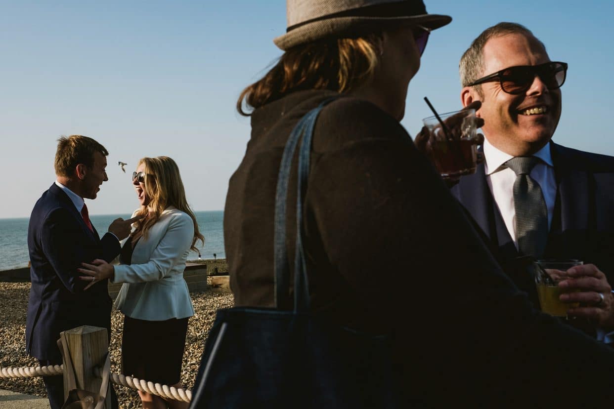 A documentary wedding photograph on the shingle beach at East Quay, Whitstable. The frame captures a group of guests chatting and interacting in an unposed, candid moment. The shot focuses on the natural human energy and the expansive, soft light of the North Sea coastline, documenting the honest atmosphere of the day.