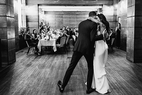 A black and white documentary photograph of a first dance at the Town Hall Hotel. The monochrome edit emphasizes the dramatic shadows, architectural textures, and the raw, timeless energy of the celebration in the Bethnal Hall.
