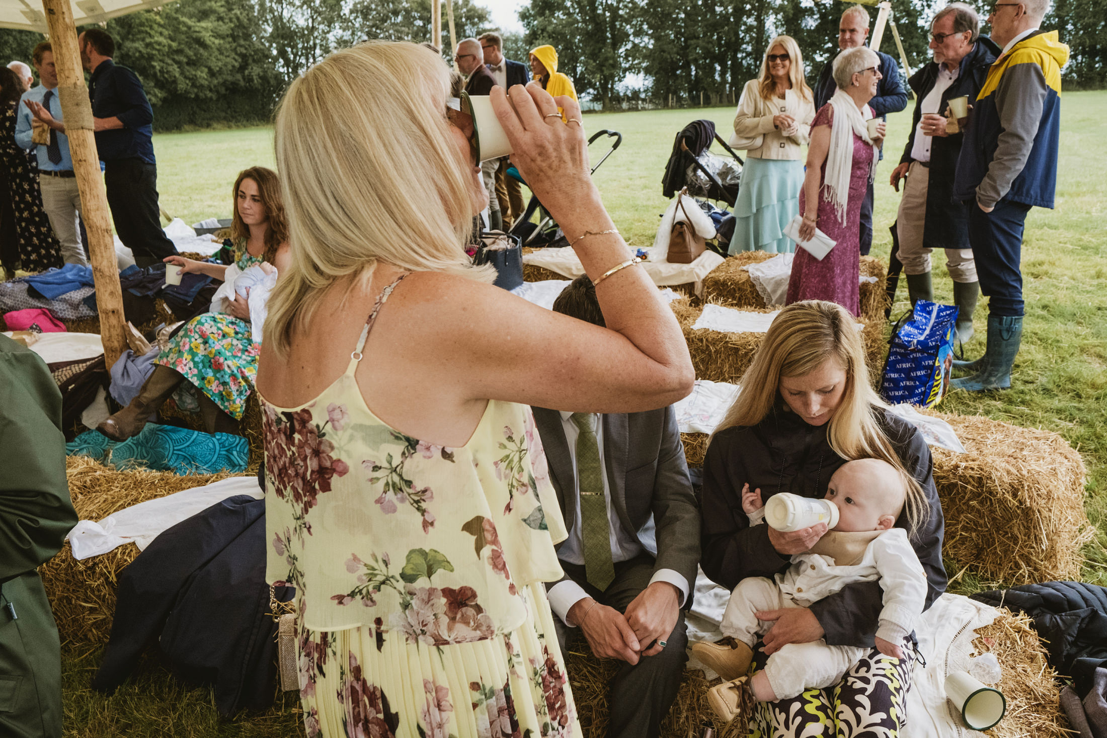 People socialising outdoors on hay bales, countryside backdrop.