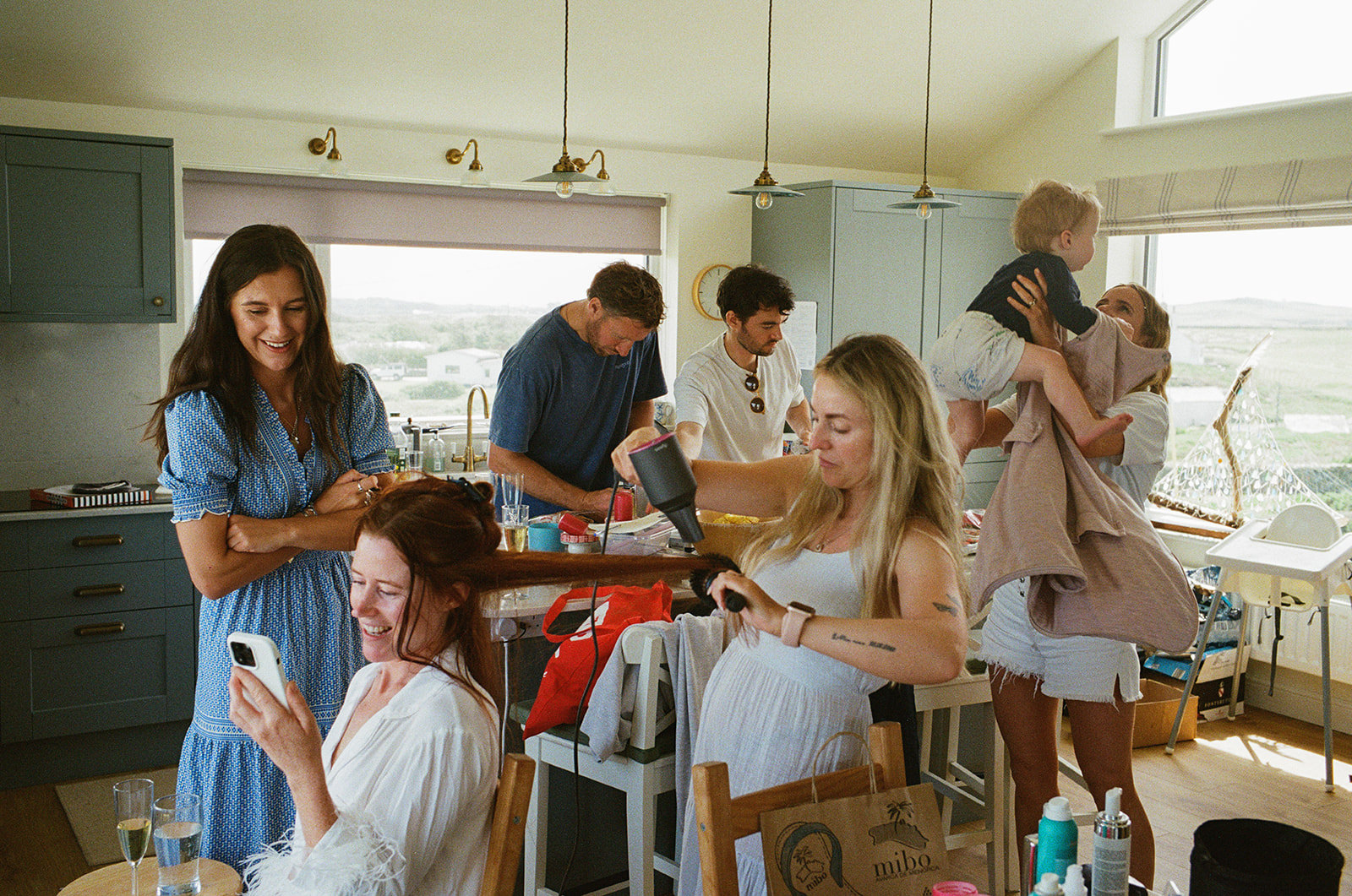 Bridal party laughing in kitchen on wedding morning as hair is styled and child lifted by window
