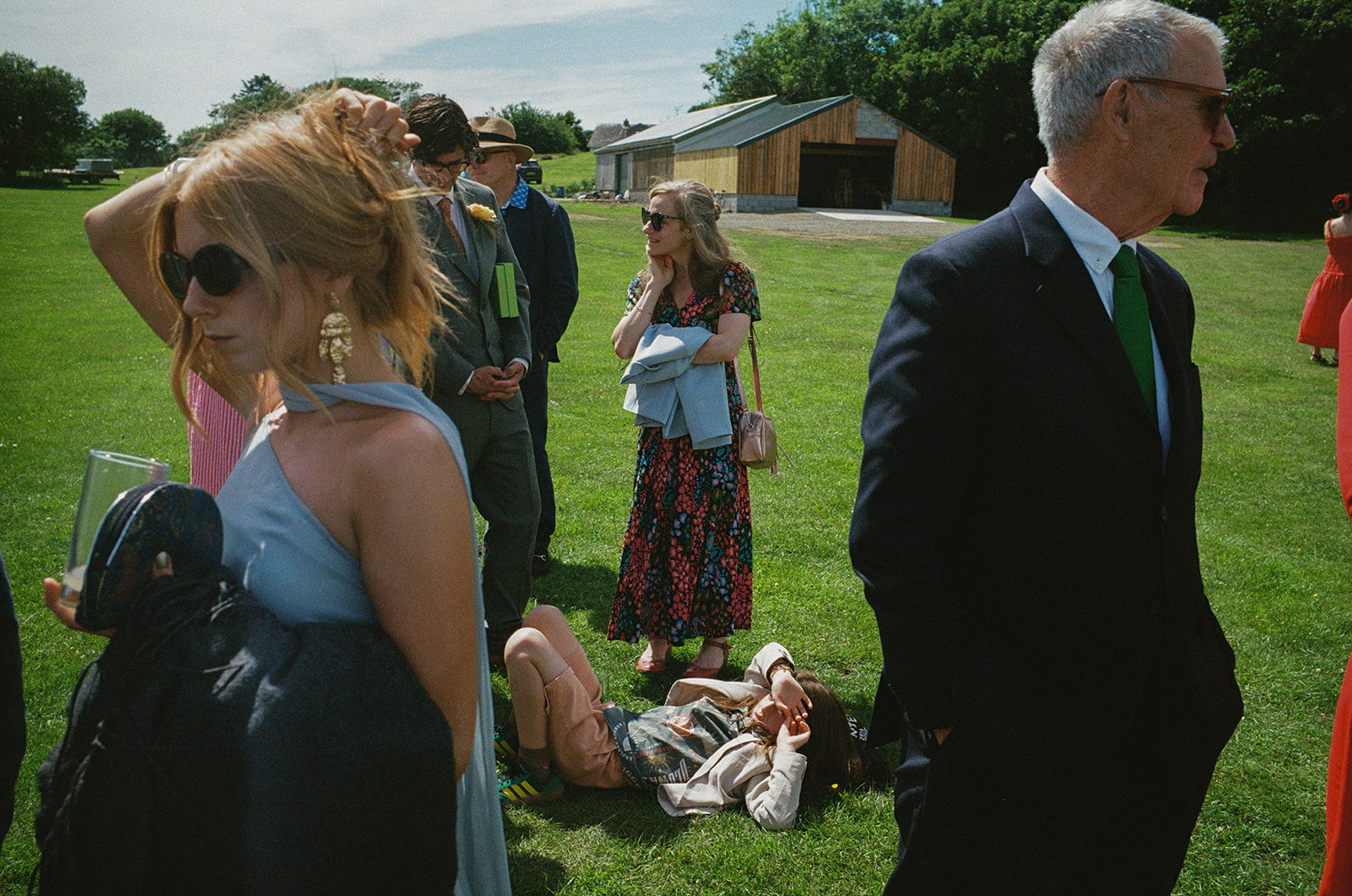 Wedding guests chat on sunlit lawn as child lies on grass near rustic barn.