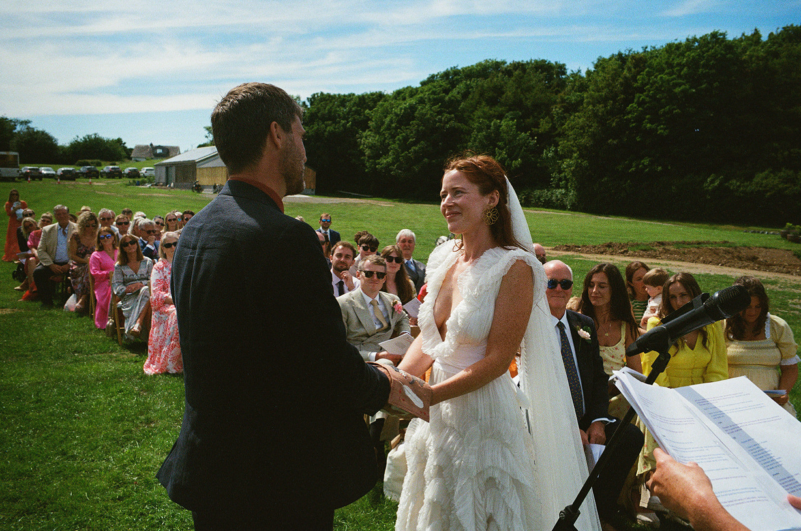 Bride and groom hold hands during outdoor wedding ceremony, guests watching in summer sunshine.
