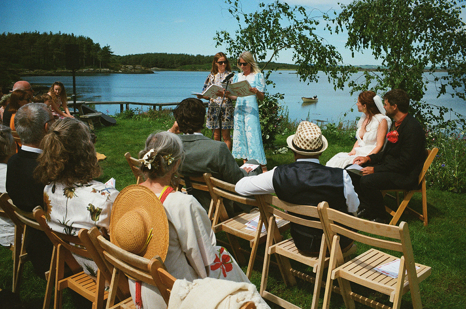 Guests seated on lawn as two women read beside a quiet lakeside wedding ceremony.