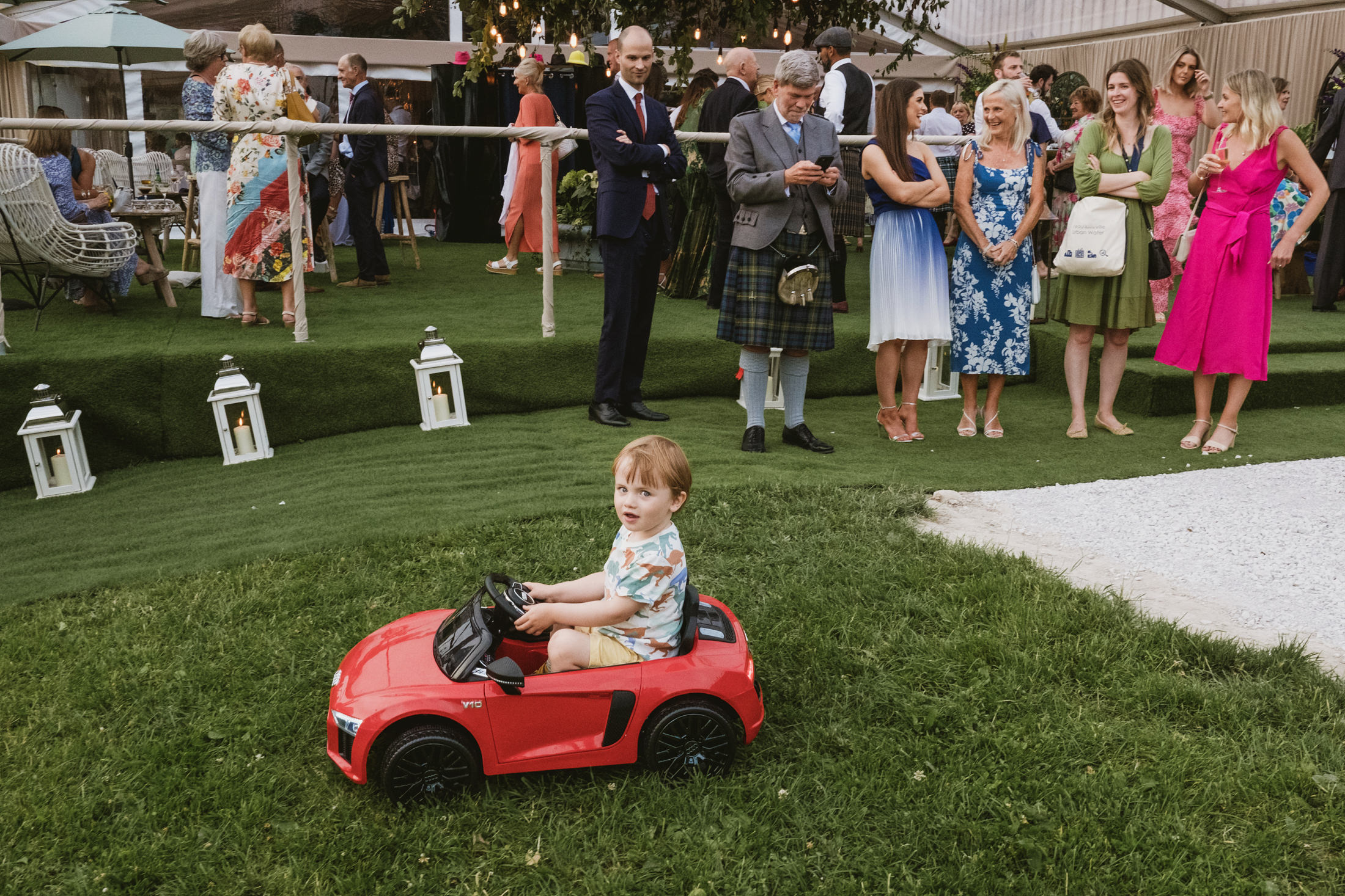 Child in toy car at garden party. Unposed and vibrant; capturing the essence of a home wedding in Denbighshire.
