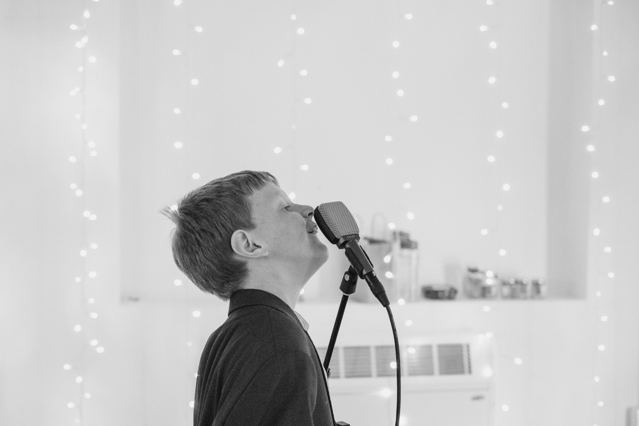 Child singing into microphone with fairy lights background.