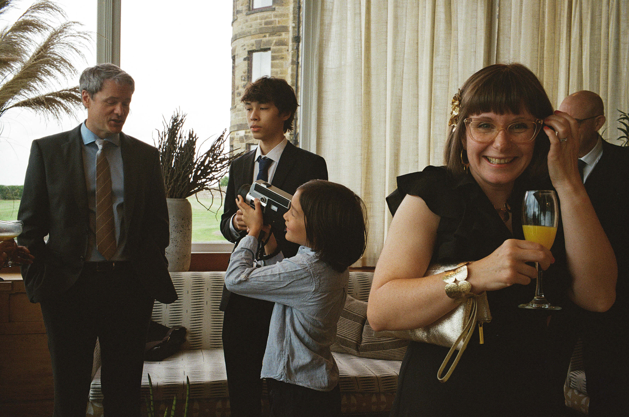 Wedding photography captured on real analogue film. Guests chatting by tall windows as child lifts camera and woman smiles with drink at wedding reception.
